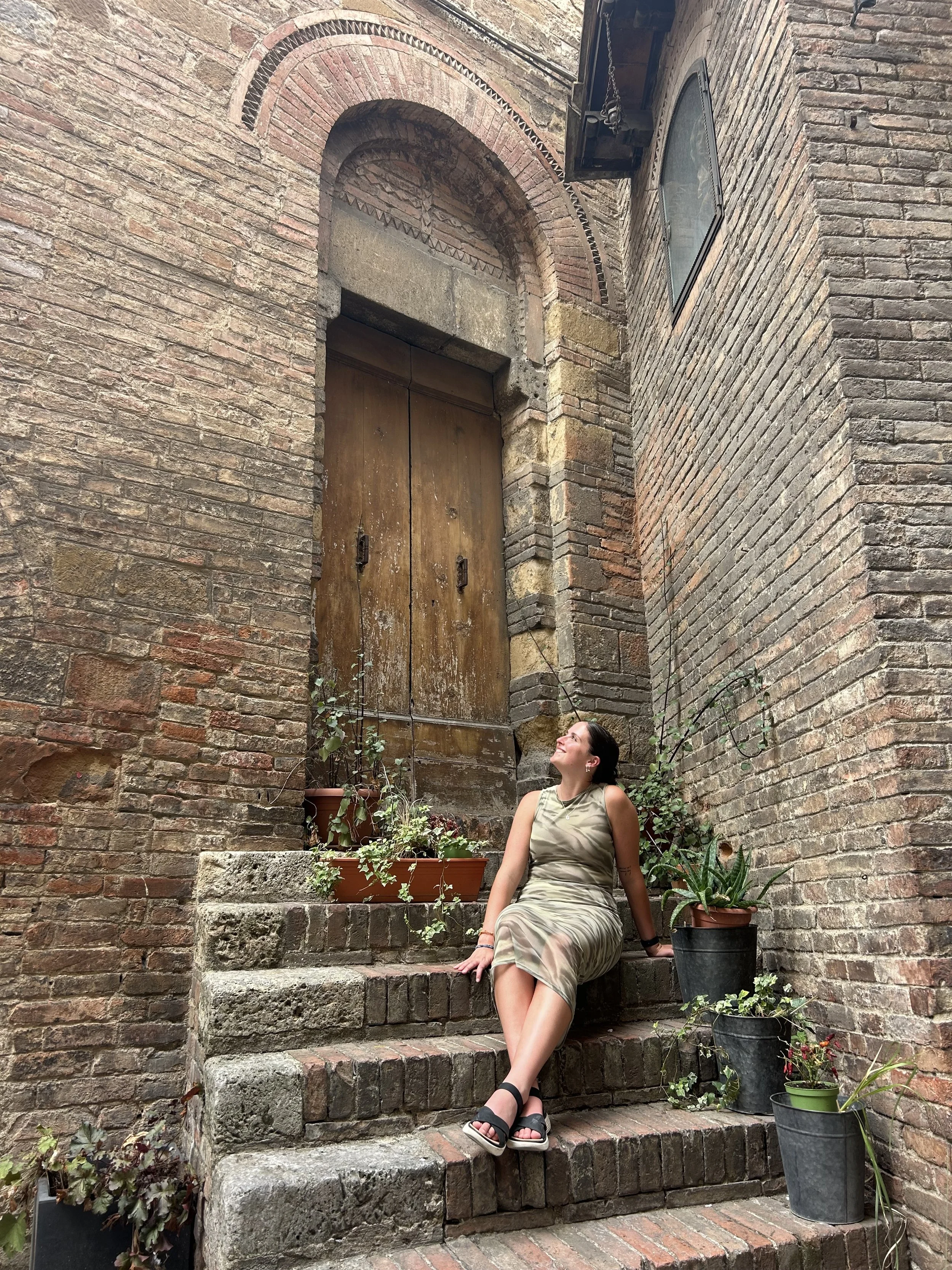 A woman in a beige dress and black sandals is sitting on stone steps next to an alley brick wall with potted plants around her, looking up at a large wooden door surrounded by brick arch and wall.