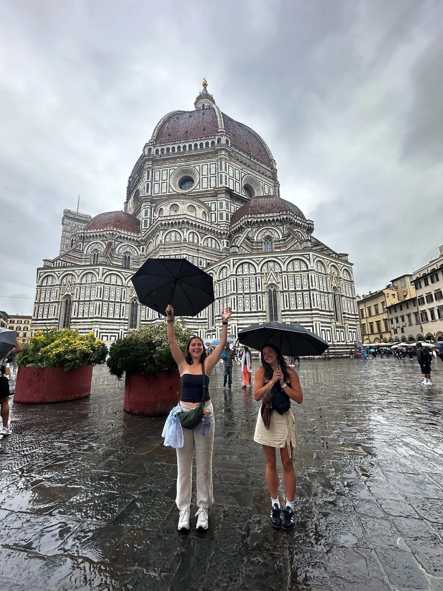 Two women are standing in front of the Florence Cathedral, holding umbrellas on a rainy day. One woman is smiling and raising her umbrella, while the other is smiling with her umbrella closed. The cathedral's building is in the background, with a cloudy sky overhead.
