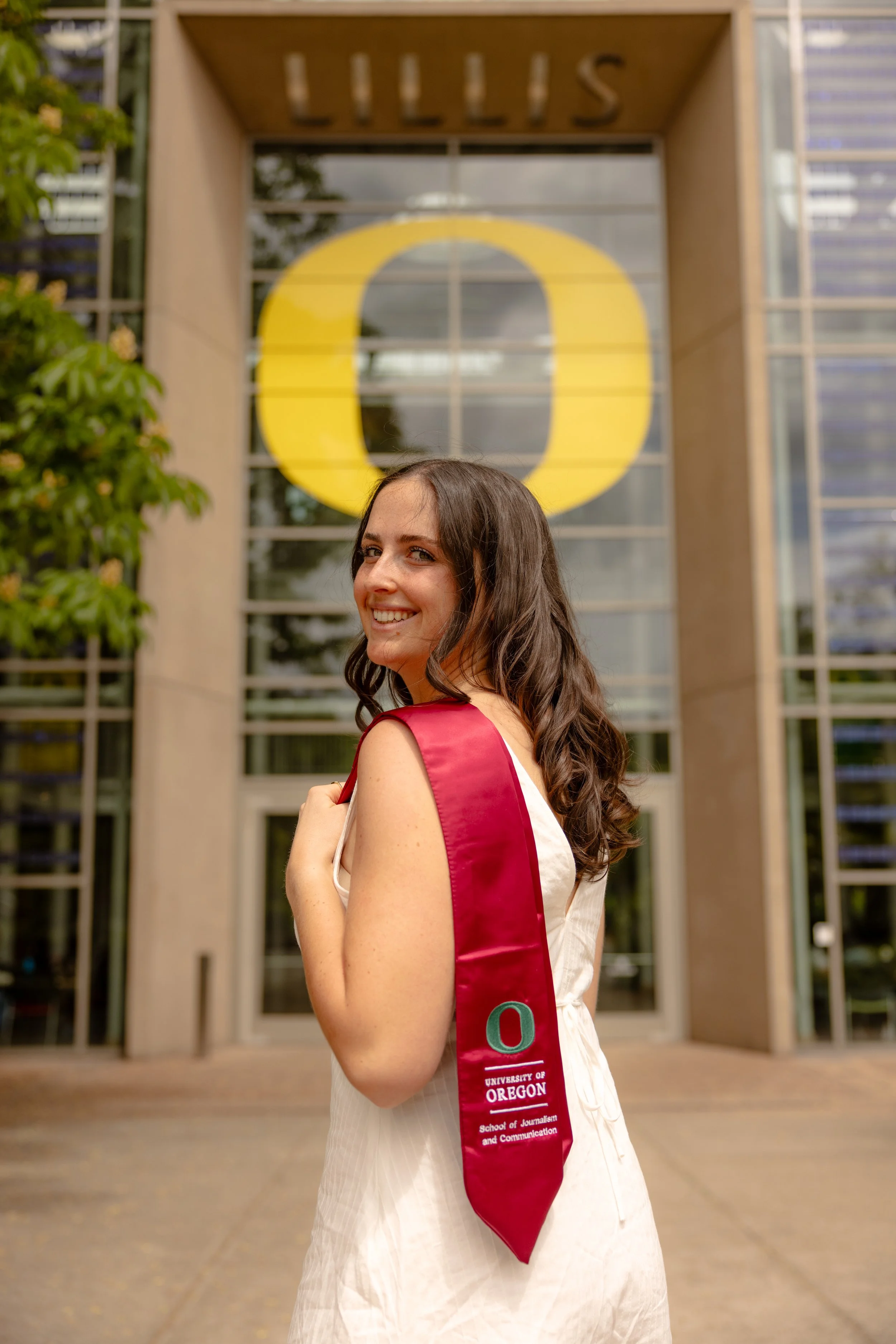 A young woman in a white dress with a red graduation sash stands in front of a modern building with glass and steel architecture, holding the sash and smiling at the camera.