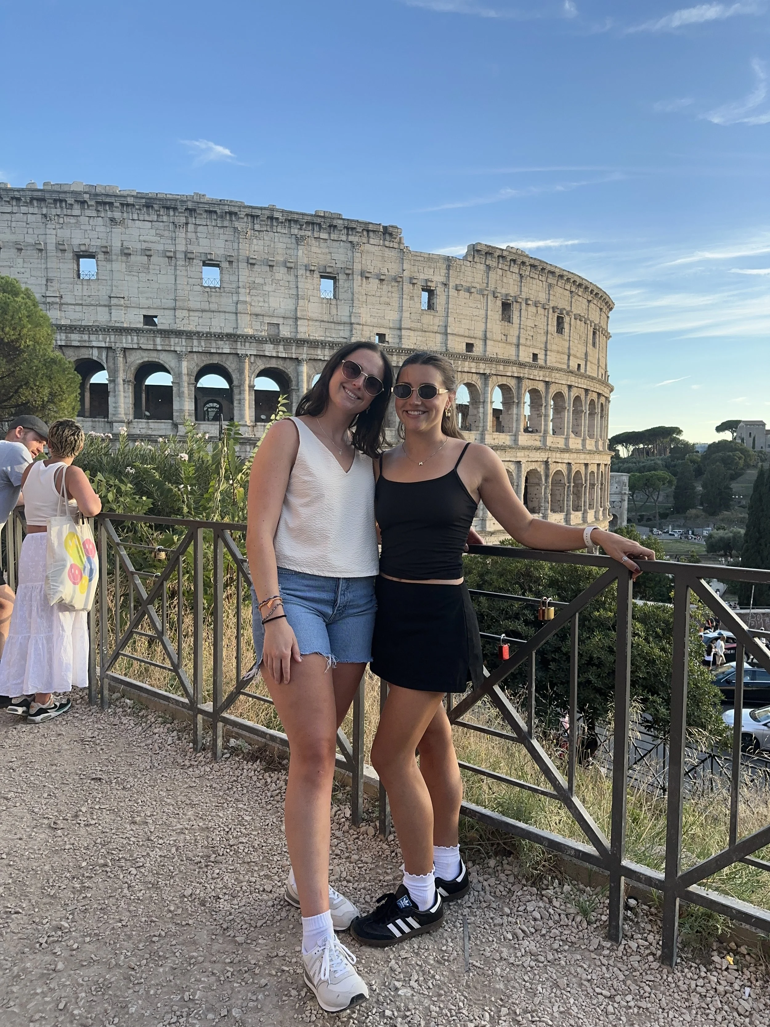 Two young women wearing sunglasses standing close together and smiling in front of the Colosseum in Rome, Italy, during daytime with a blue sky and few clouds.