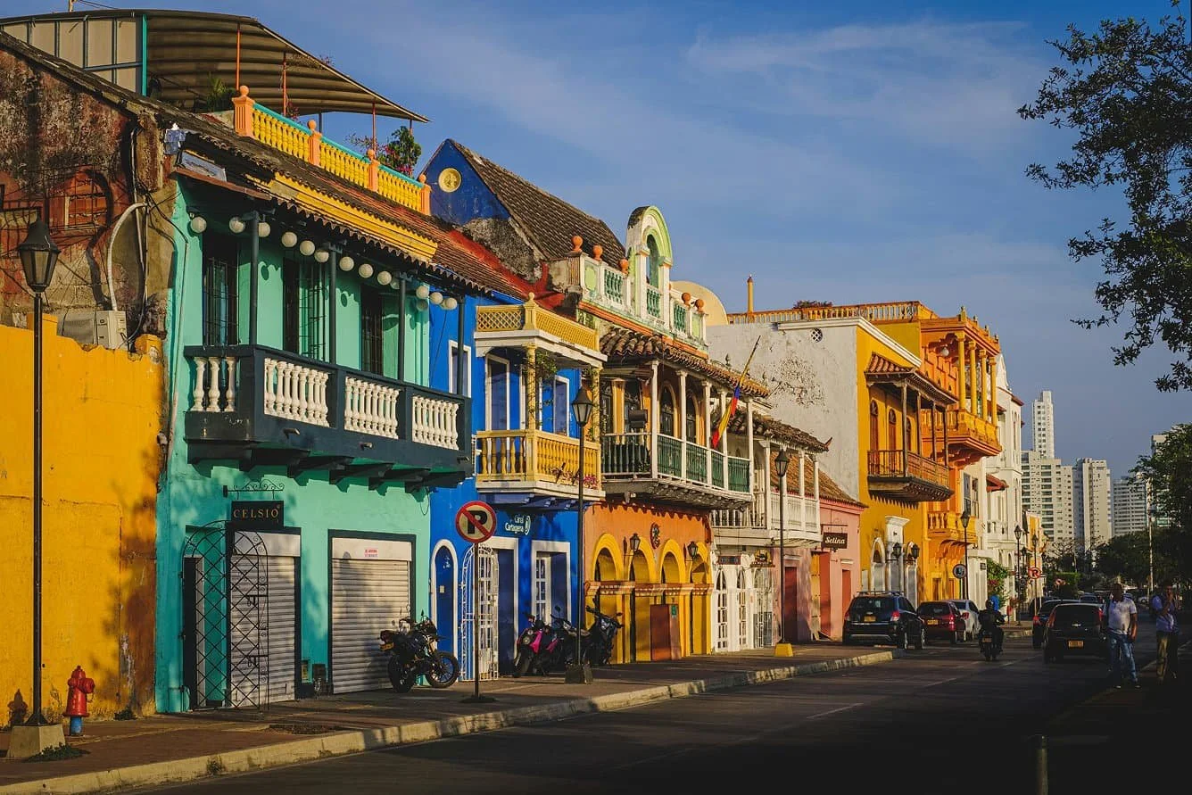 Una calle soleada está flanqueada por coloridos edificios coloniales con balcones; a lo largo de la calle hay coches y motos aparcados.