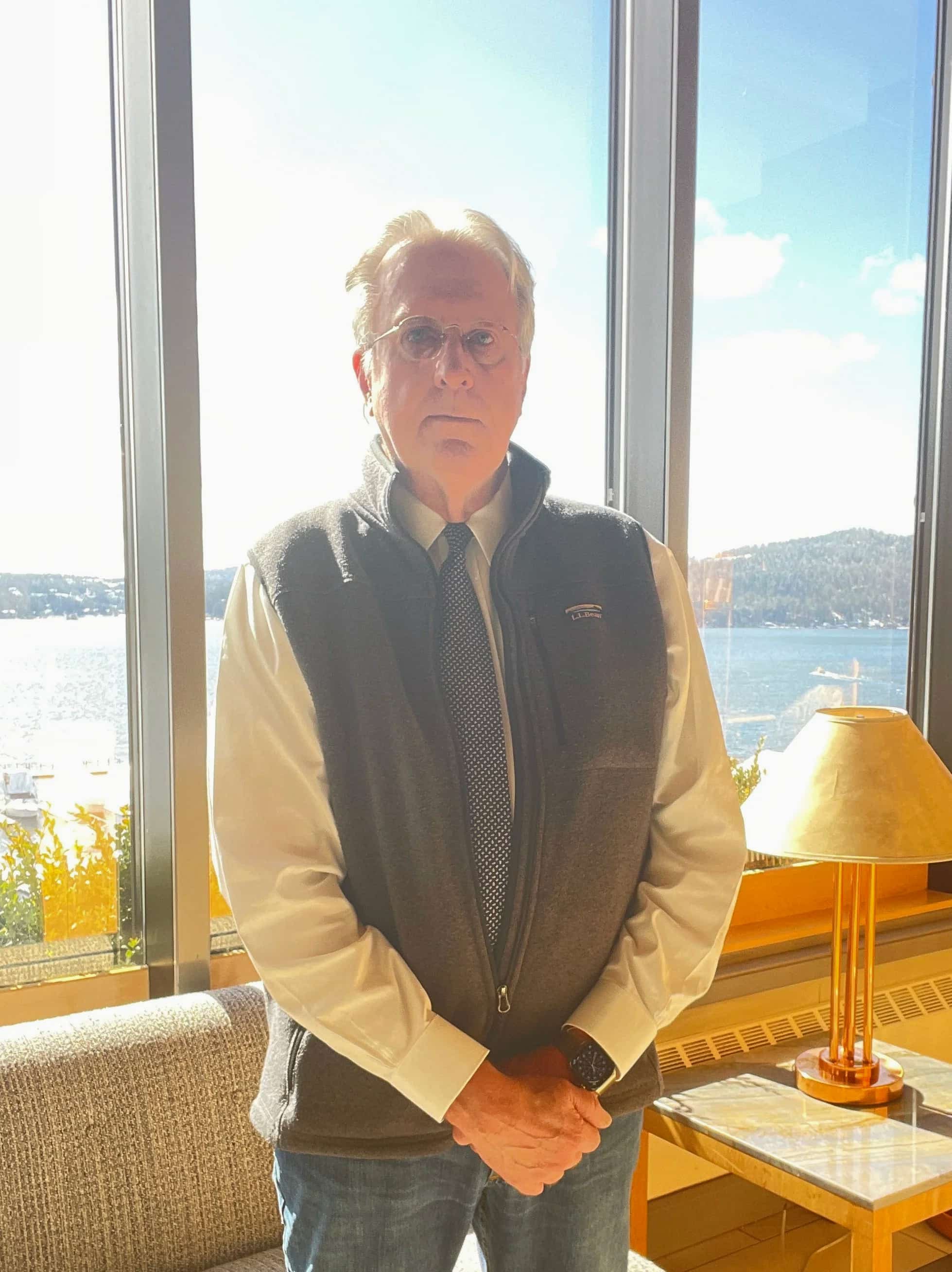 Un hombre mayor con gafas y corbata está de pie en el interior, junto a unos grandes ventanales, con la luz del sol y una pintoresca vista del lago al fondo.