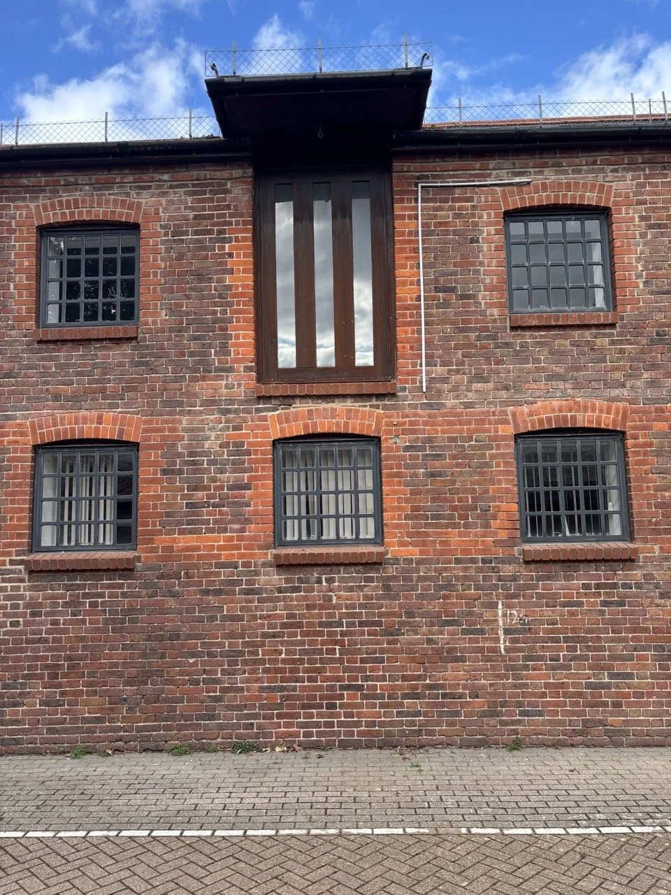 Brick building with six windows, five with black grids and one large vertical window, under a cloudy sky.