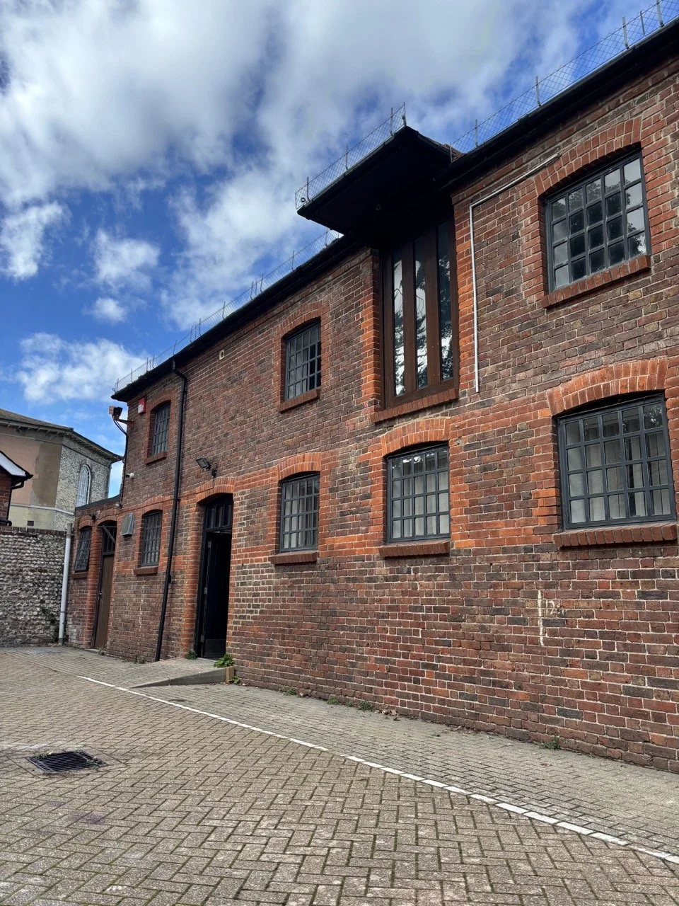 A brick building with black window frames and a black door, surrounded by a paved courtyard, under a partly cloudy sky.