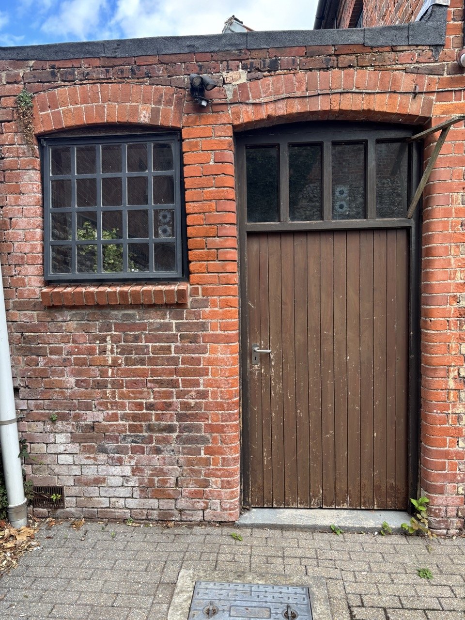 Brick building exterior with a brown wooden door, a black metal framed window, security camera, and small green plants near the ground.