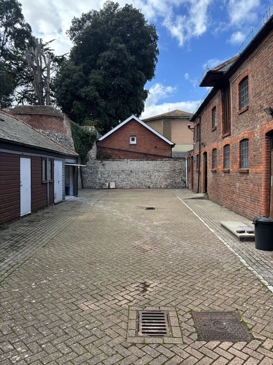 An empty brick-paved courtyard bordered by old brick buildings, with a large tree in the background and a blue sky with a few clouds.