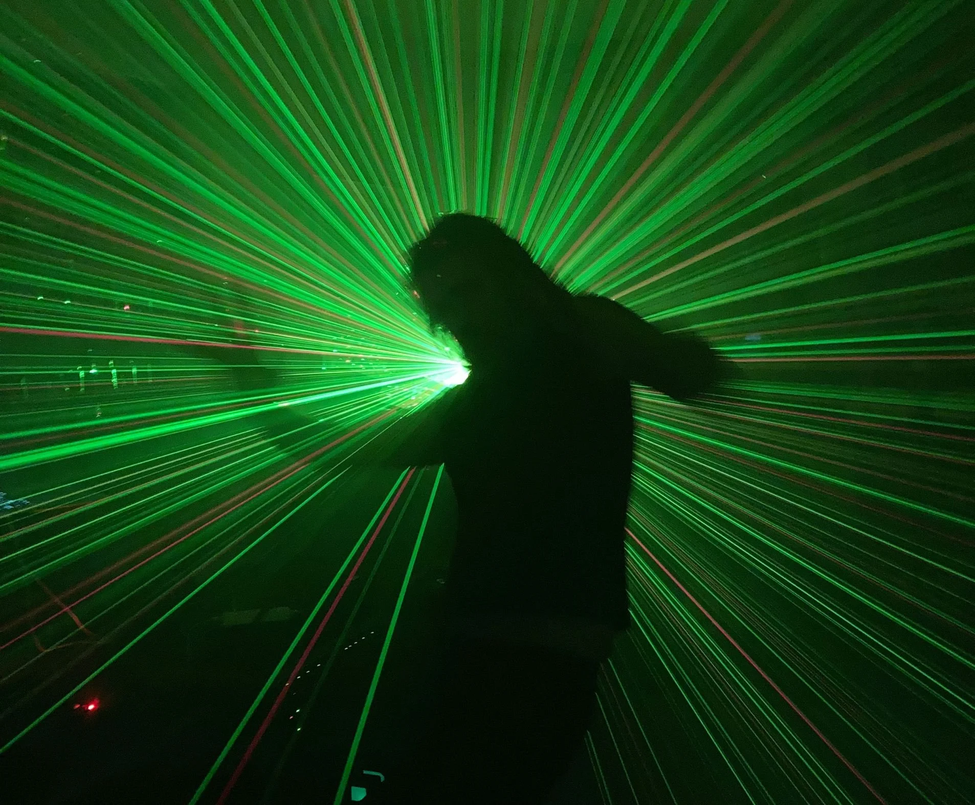 A person is holding a green laser pointer creating a pattern of green light beams radiating outward, with some red light at the bottom left corner.