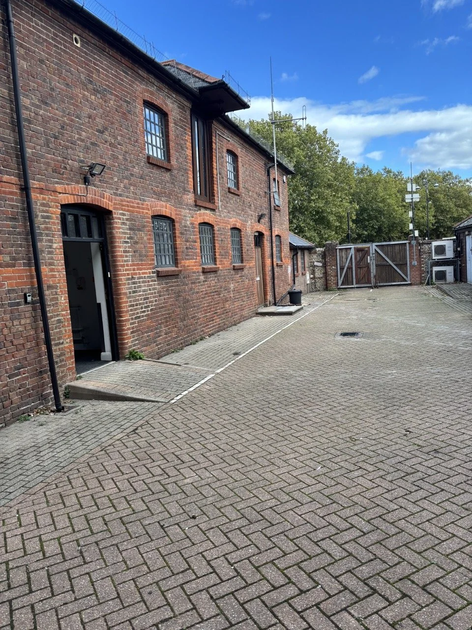 A brick building with multiple windows and a small door, situated in a paved alley with a wooden gate at the end, and trees with green leaves in the background.