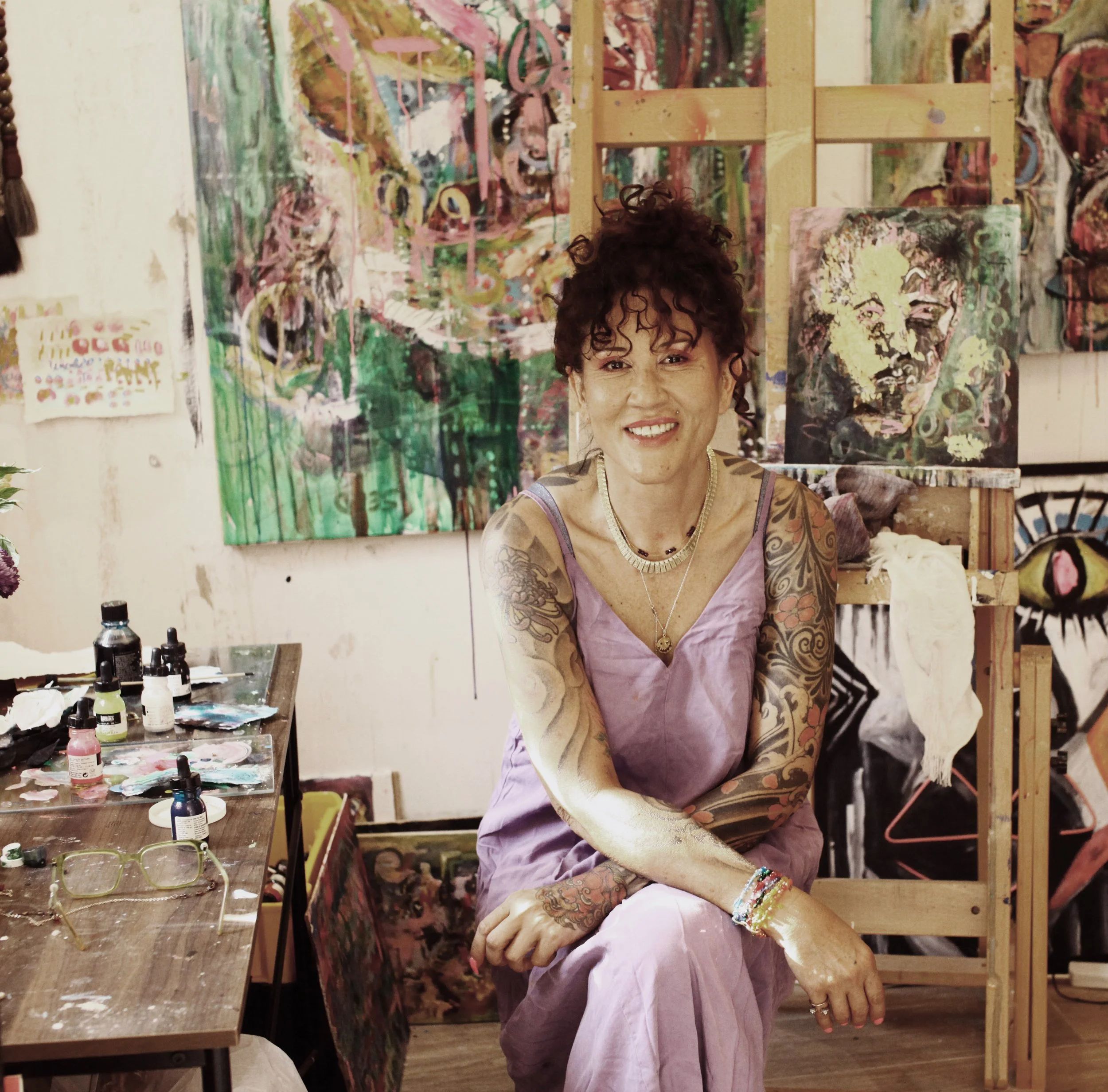A person with curly hair, tattoos, and jewelry is smiling in an art studio. The background displays colorful, abstract paintings and art supplies on a table.