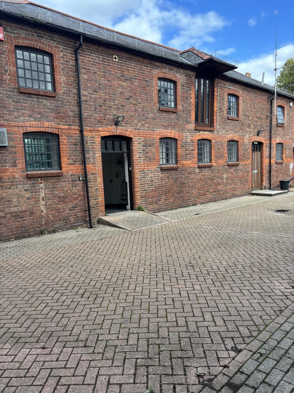 Red brick building with multiple black-framed windows, a black door, and a paved courtyard area under a partly cloudy sky.