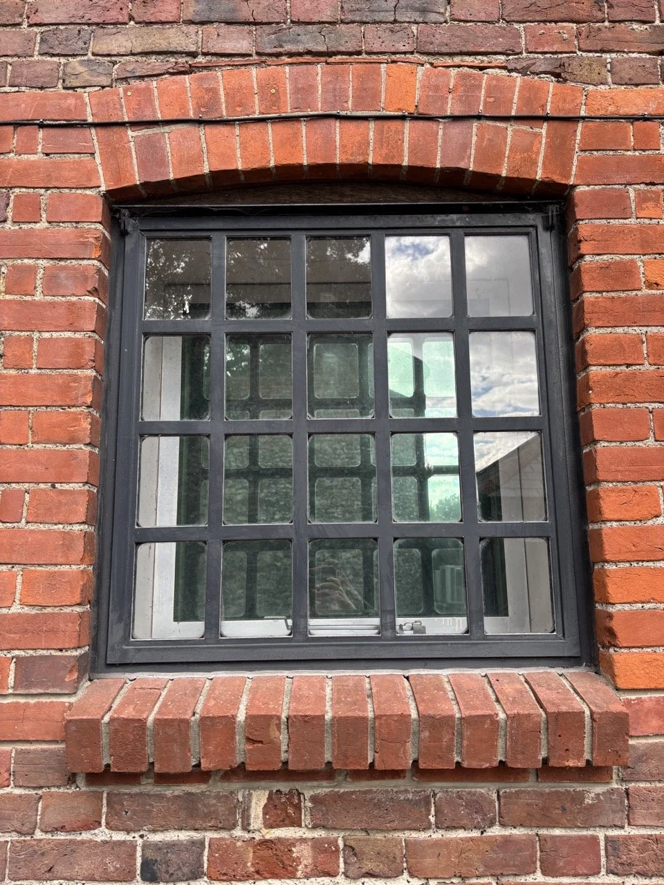 A square window with a black metal grid, set in a red brick wall, reflecting clouds and trees.