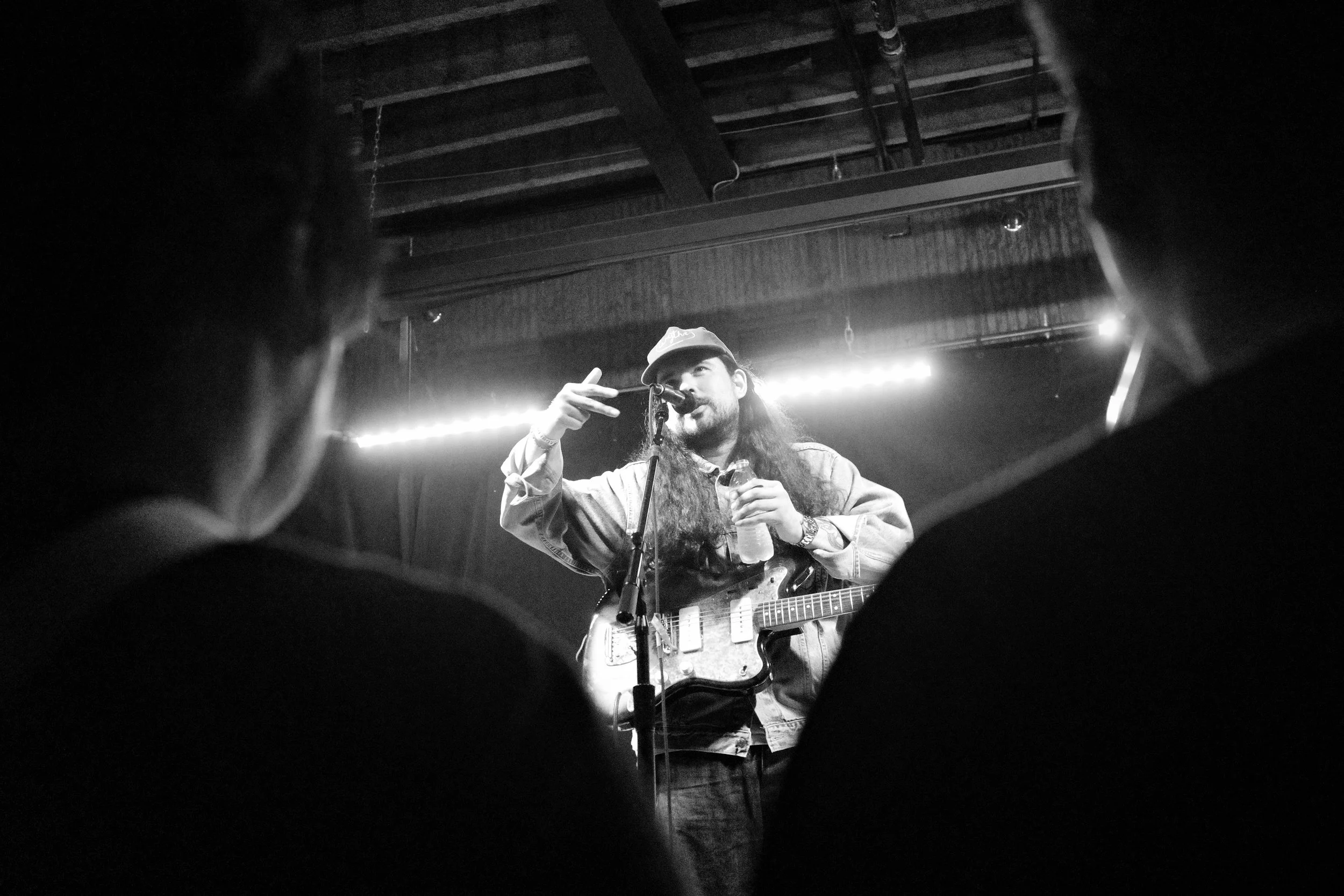 A musician with long hair and a cap on stage, holding a beverage and using a guitar, performing under stage lights, viewed from the audience.