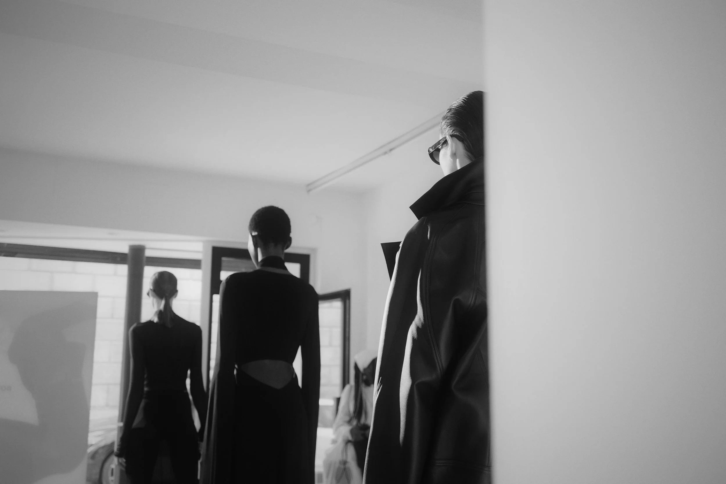 Black-and-white photo of three women from behind, wearing fashionable clothing and sunglasses, standing indoors near an open window.