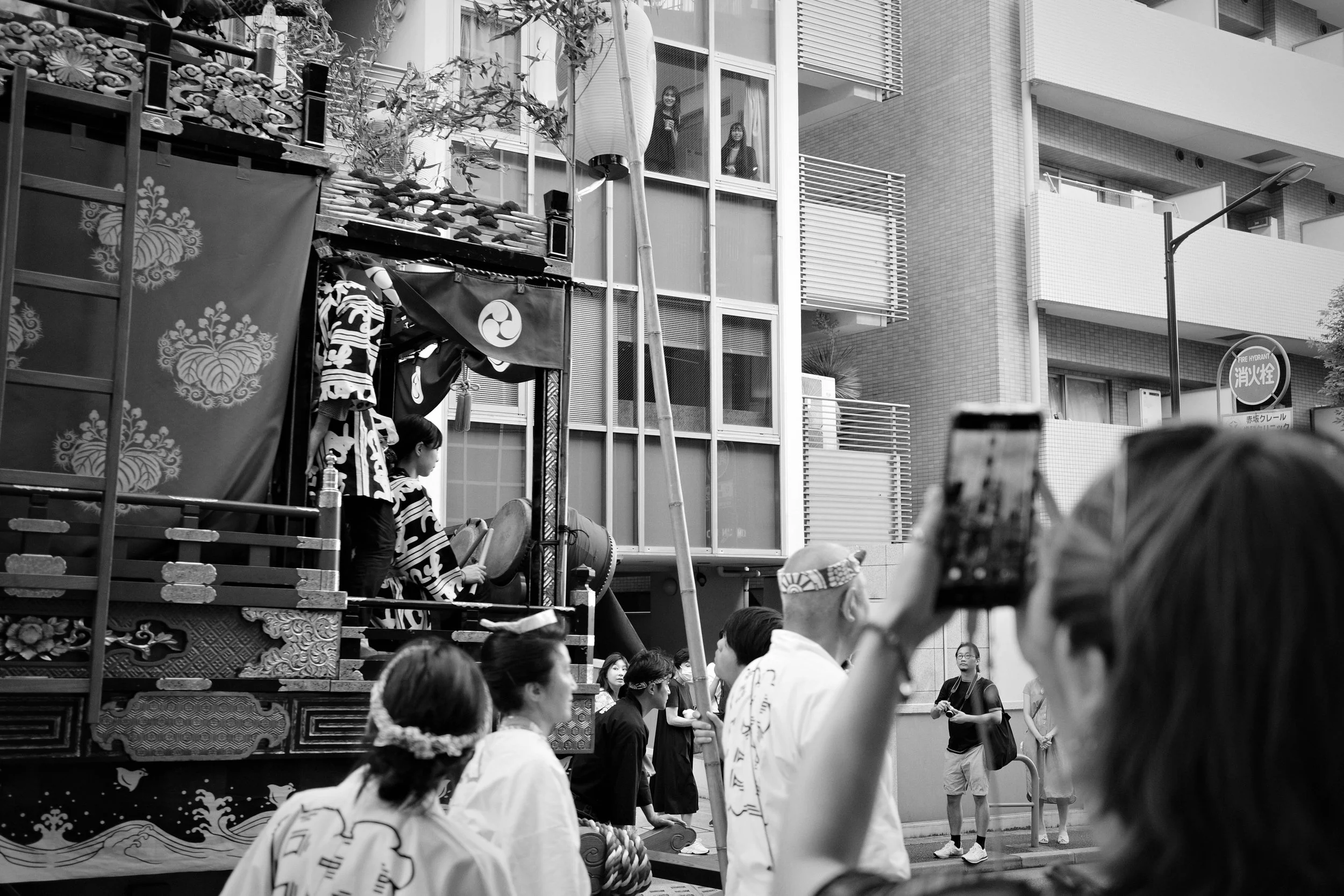 A black and white photo of a traditional Japanese festival float being paraded through a city street. People dressed in traditional attire, and some are watching and taking photos. A float has dolls or figures on it and decorative elements.