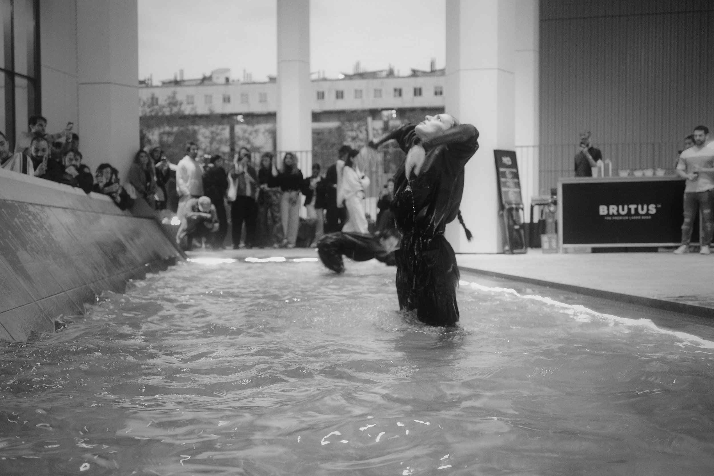 A woman is seen in a pool during a fashion show, wearing a black outfit with water dripping off her, with a crowd of people watching in the background.