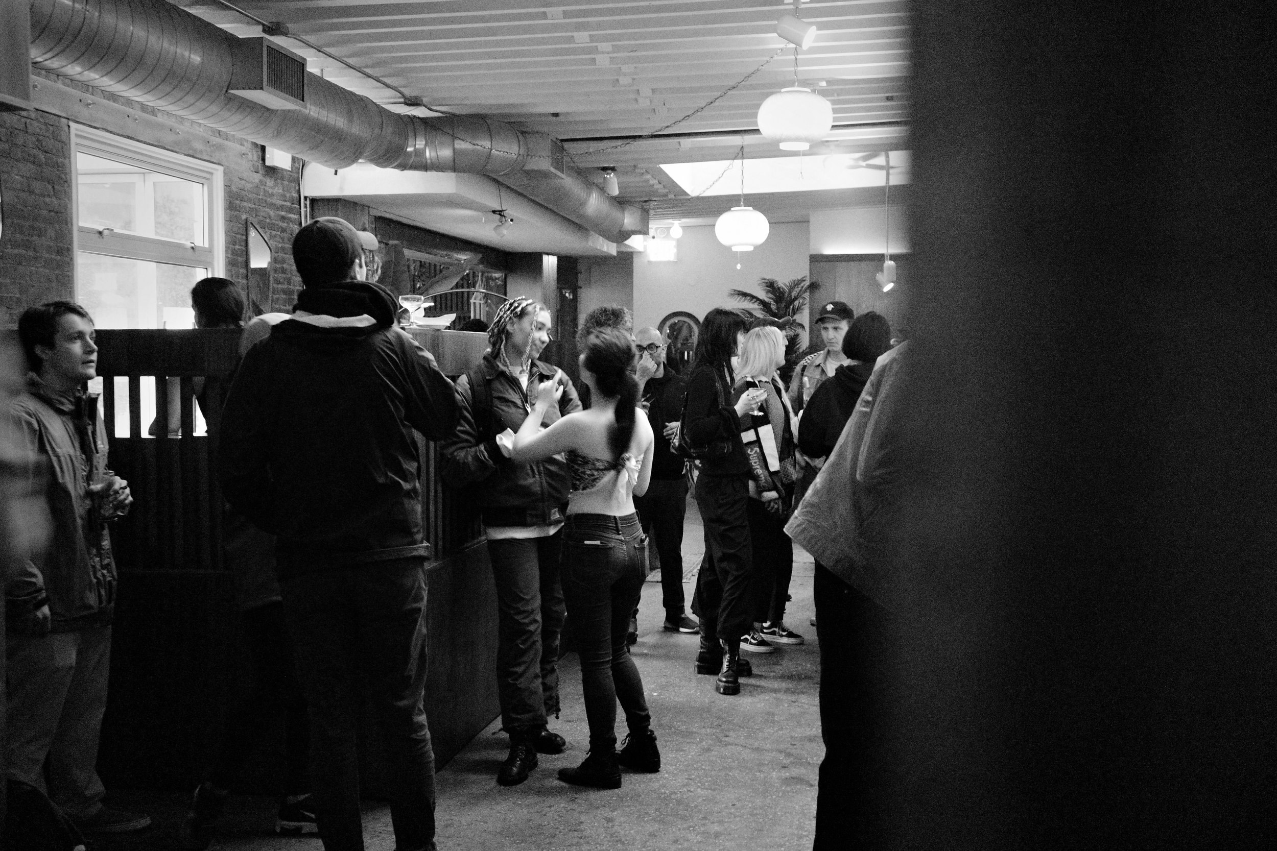 People standing in line in a bar or club, illuminated by hanging lights, with brick walls and industrial ceiling pipes in the background.
