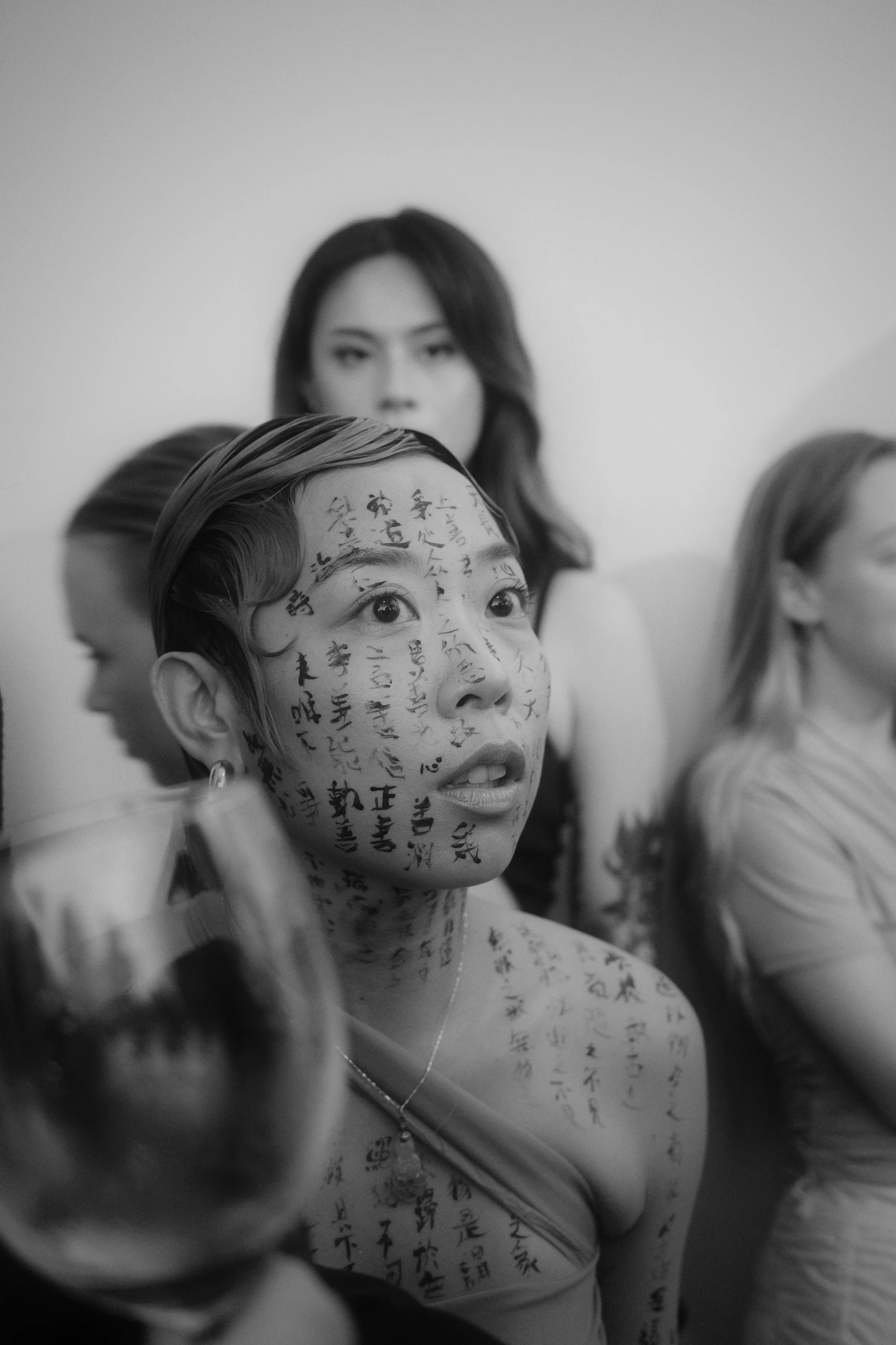 A woman with traditional Chinese writing on her face and body, sitting among other women at a gathering or event.
