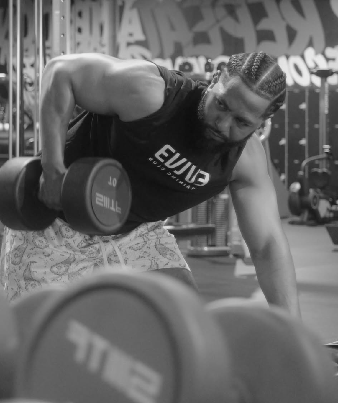 A man with braided hair and a beard performs a bent-over dumbbell row exercise in a gym, wearing a black tank top and patterned shorts.