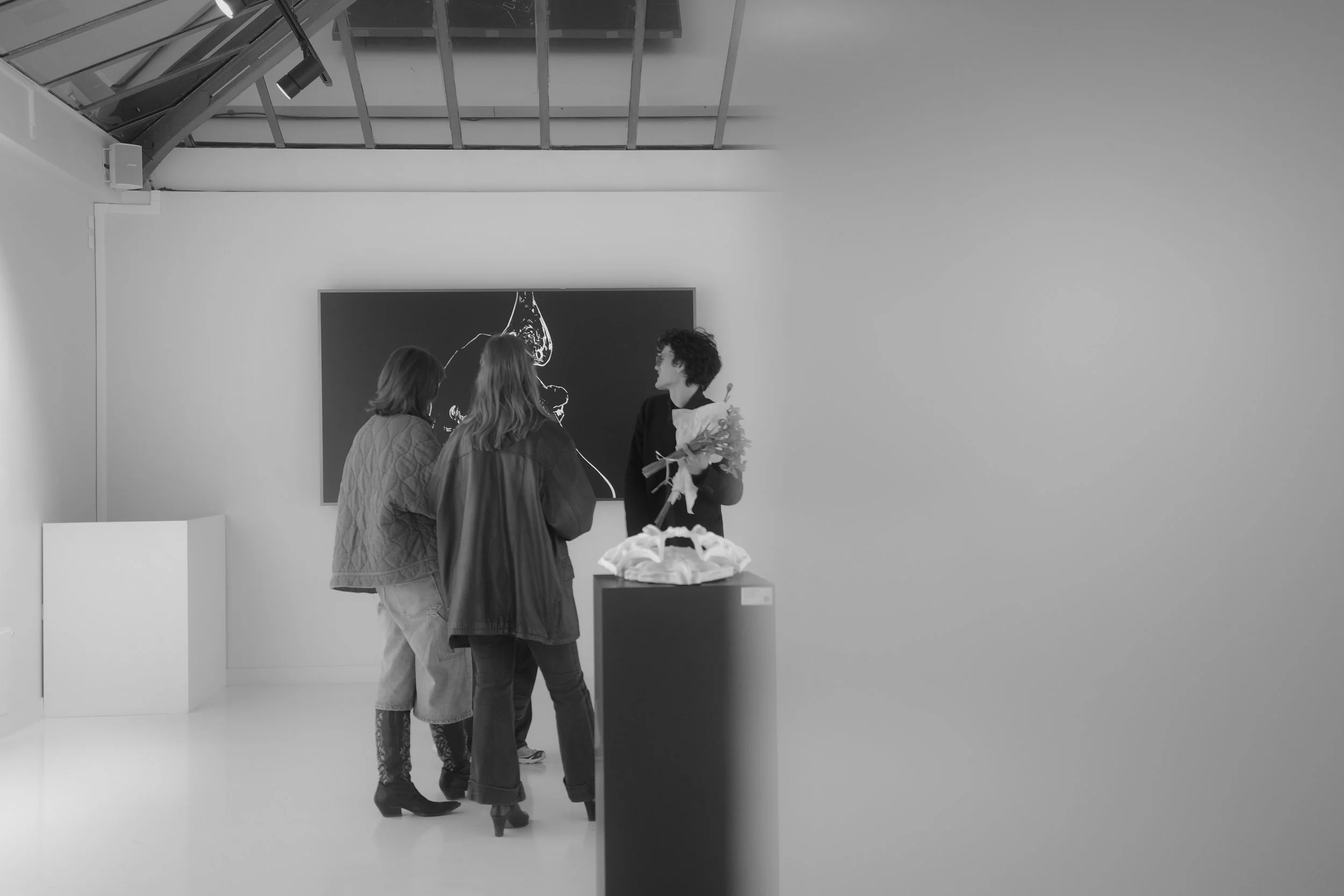 Three people are standing in an art gallery, talking. One person is holding a bouquet of flowers. The gallery features a large black and white artwork on the wall and a sculpture on a pedestal in the foreground. The ceiling has a glass roof.