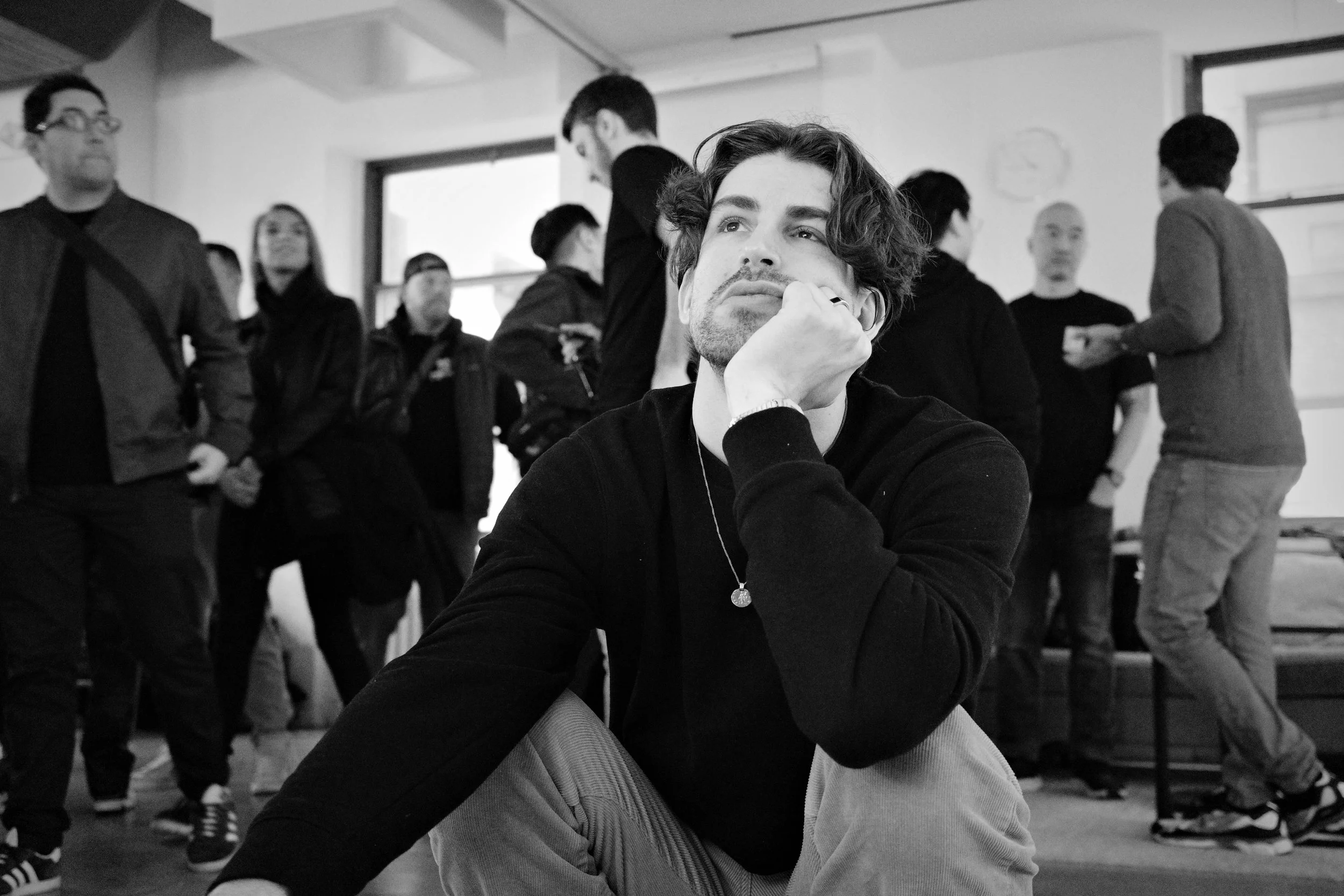 A man with wavy hair and a beard sitting on the floor with his chin resting on his hand in a busy room with several people standing and talking in the background.