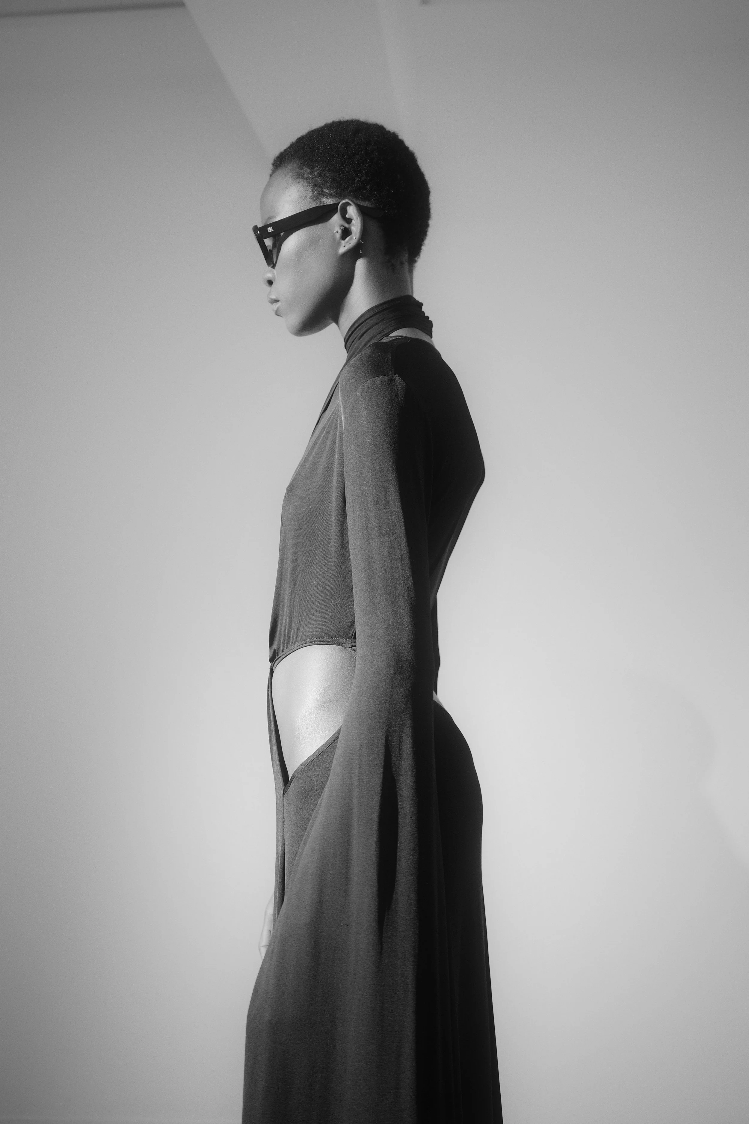 Side profile of a woman with short hair wearing sunglasses, a high-neck top, and a long coat, in monochrome.