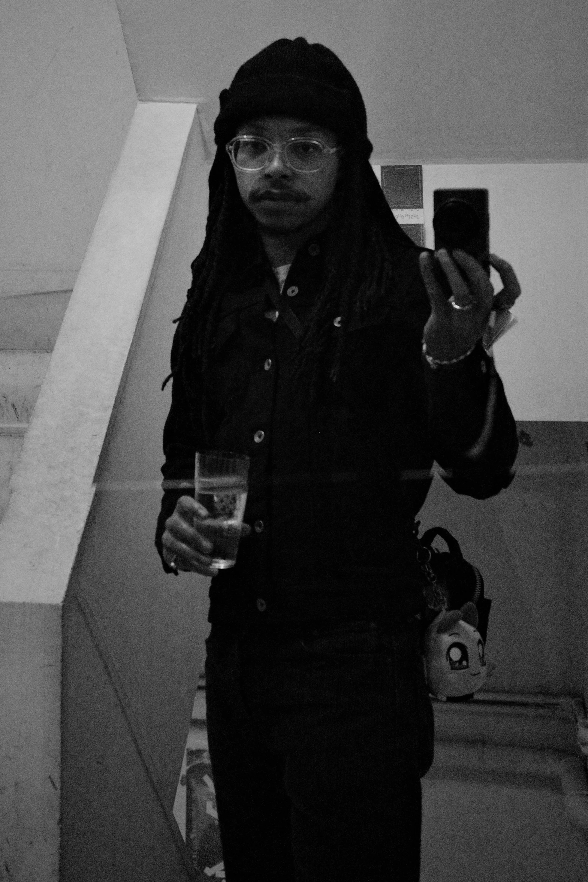 A man with long dreadlocks, wearing glasses, a beanie, and a dark jacket, is taking a mirror selfie in black and white. He is holding a glass of water in his left hand and a phone in his right hand. There is a stuffed toy on a shelf in the background.