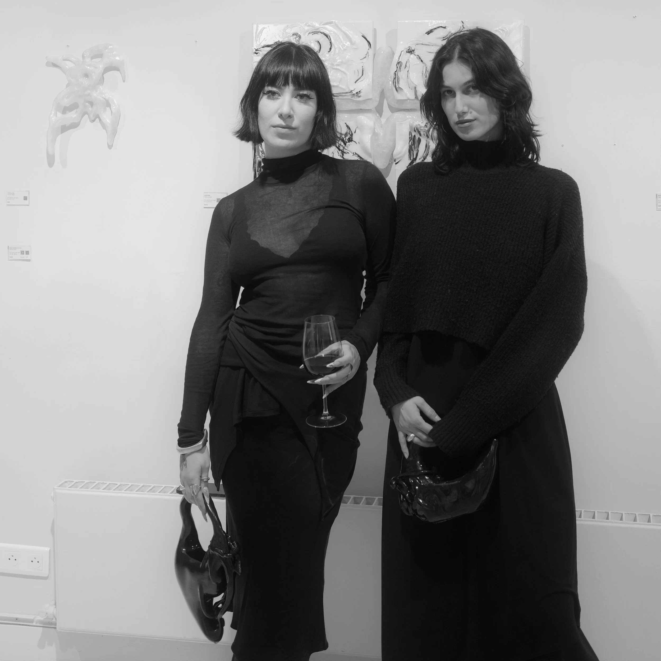 Two women standing side by side in an art gallery, both dressed in black, holding black handbags, with artwork on the wall behind them.