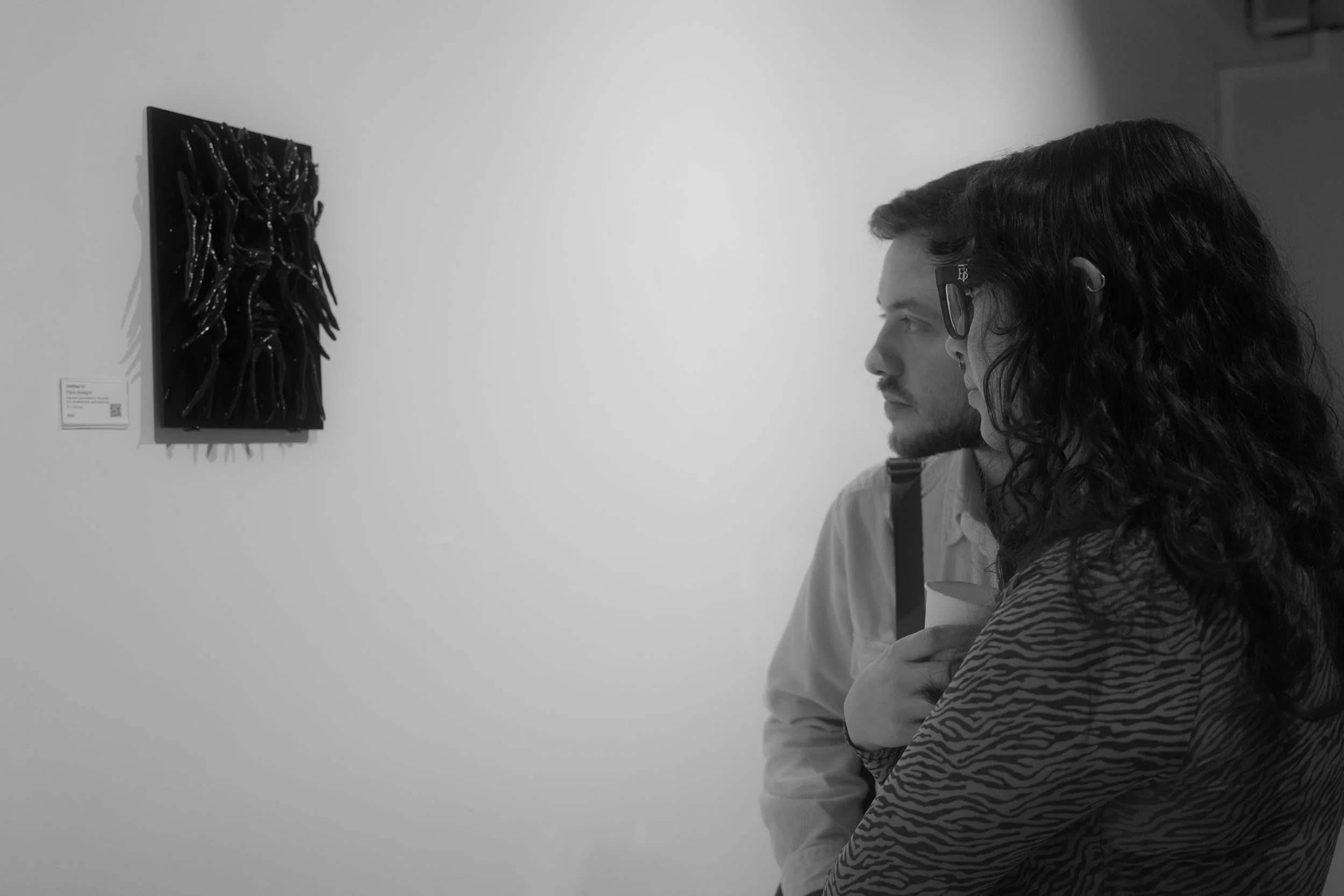 Two people observing an artwork in an art gallery. The artwork appears to be a textured, multi-layered piece with dark, shiny elements. One person has long, wavy hair and glasses, while the other has short hair and a beard.