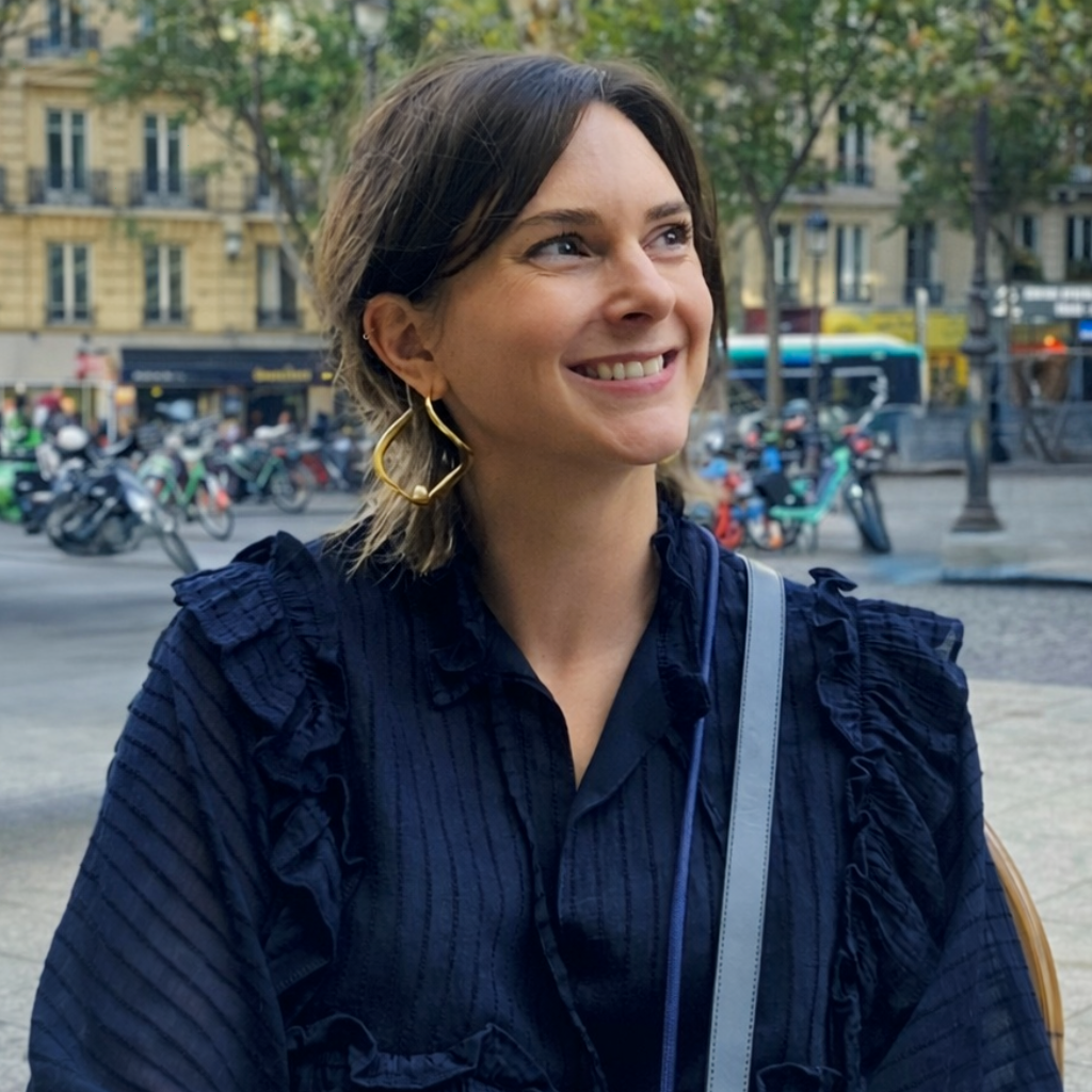 A smiling woman with short brown hair wearing a navy blue ruffled blouse, large gold earrings, and a shoulder bag, standing outdoors in an urban city setting with trees and buildings in the background.
