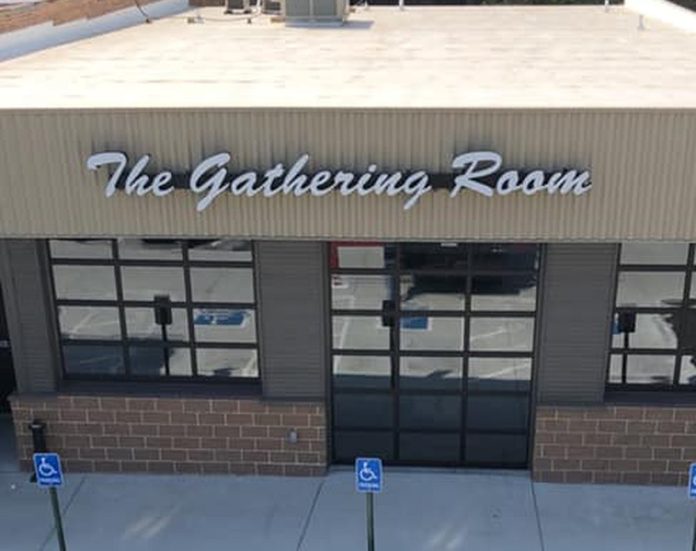Exterior of a building with a sign that reads 'The Gathering Room,' featuring large windows and accessible parking spaces.