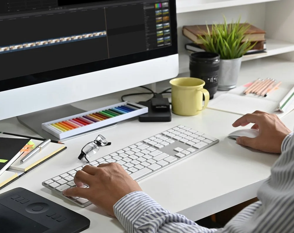 A person working on a computer at a cluttered white desk with a keyboard, mouse, glasses, pens, a yellow mug, a box of colored chalk, notebooks, a camera lens, a potted plant, books, and pencils.