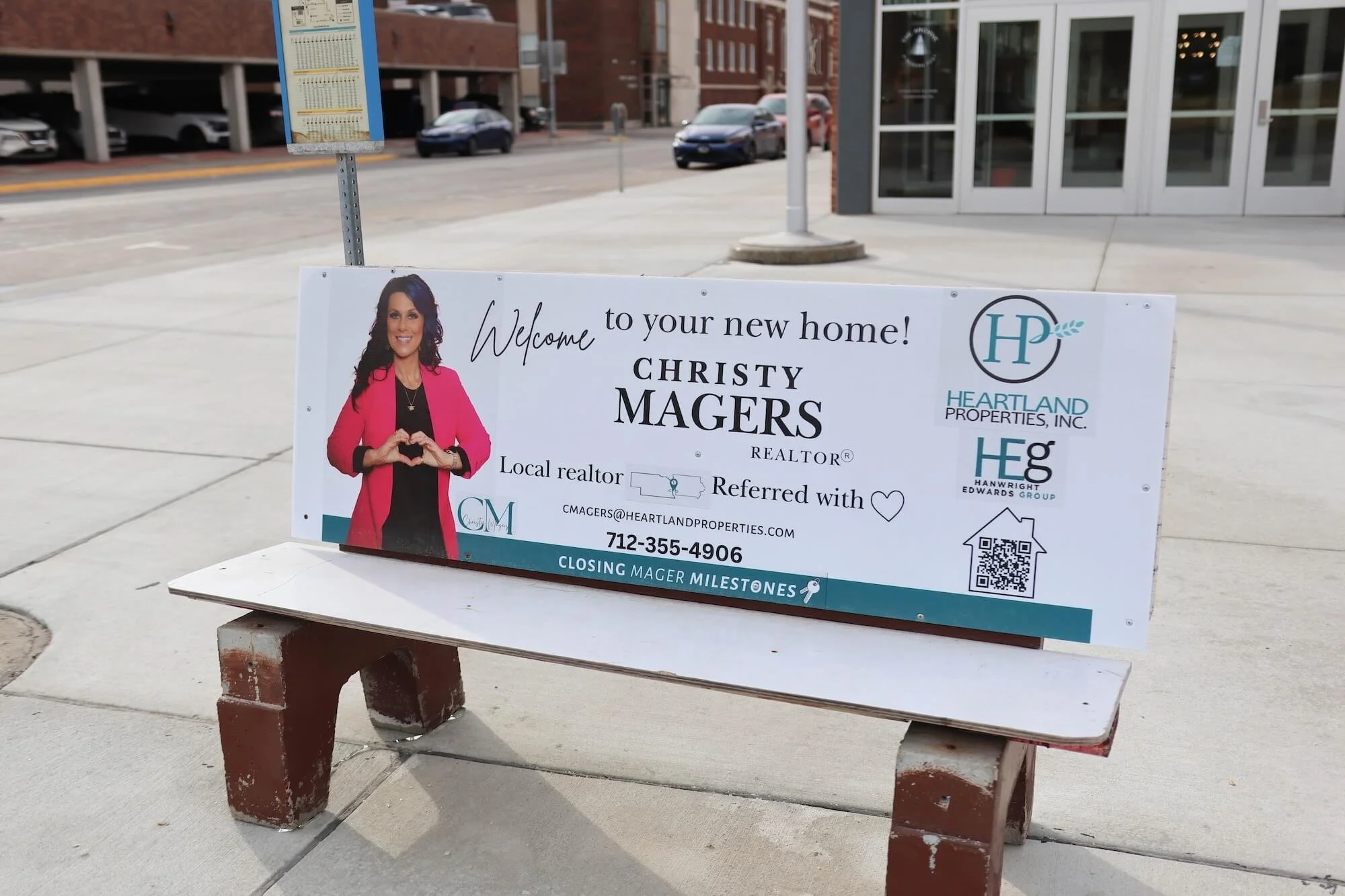 A welcome sign for Christy Magers, a realtor, displayed outside on a bench, featuring her photo making a heart gesture with her hands, with contact information and logos of real estate companies, near a building and a sidewalk.