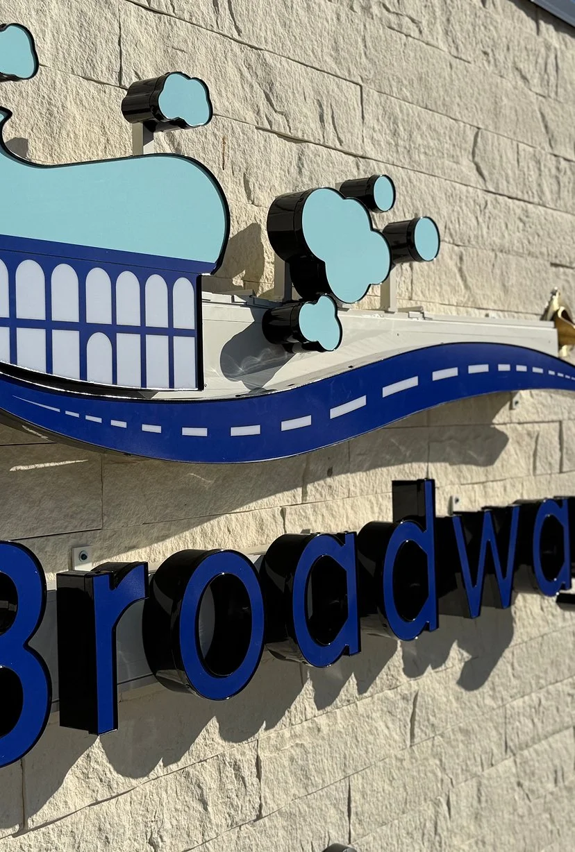 Close-up of a sign with the word "Broadway" and a decorative graphic featuring stylized clouds and a curved road on a beige brick wall.