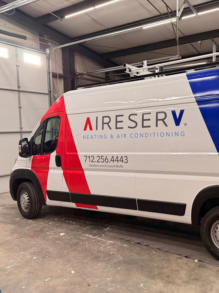 A delivery van inside a garage with branding for AireServ Heating & Air Conditioning, featuring red, white, and blue colors, a phone number, and a website link.