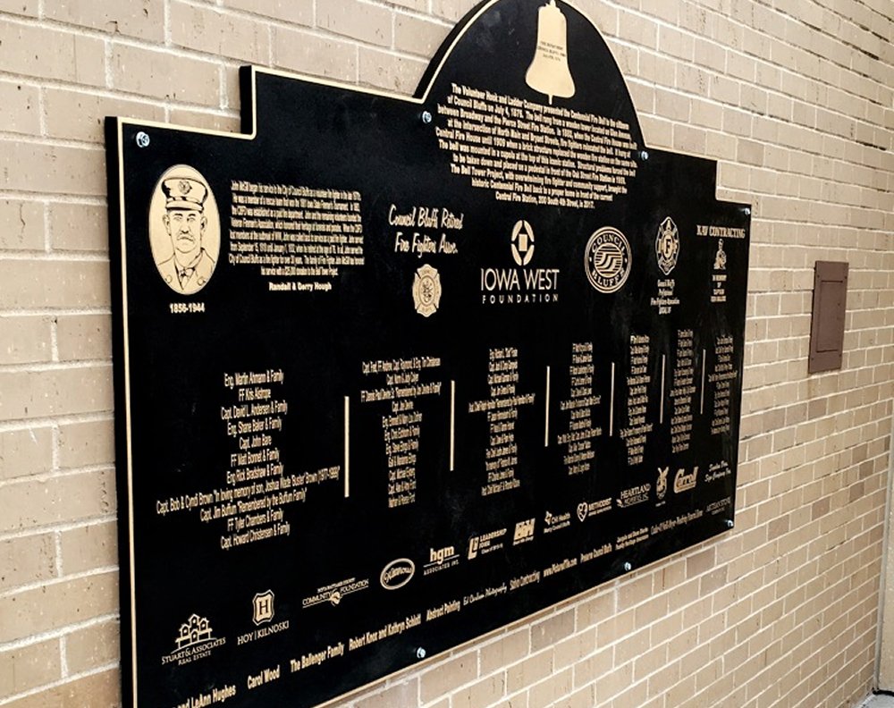 Black memorial plaque on a brick wall with text and logos, including a cartoon portrait of a man in a military uniform.