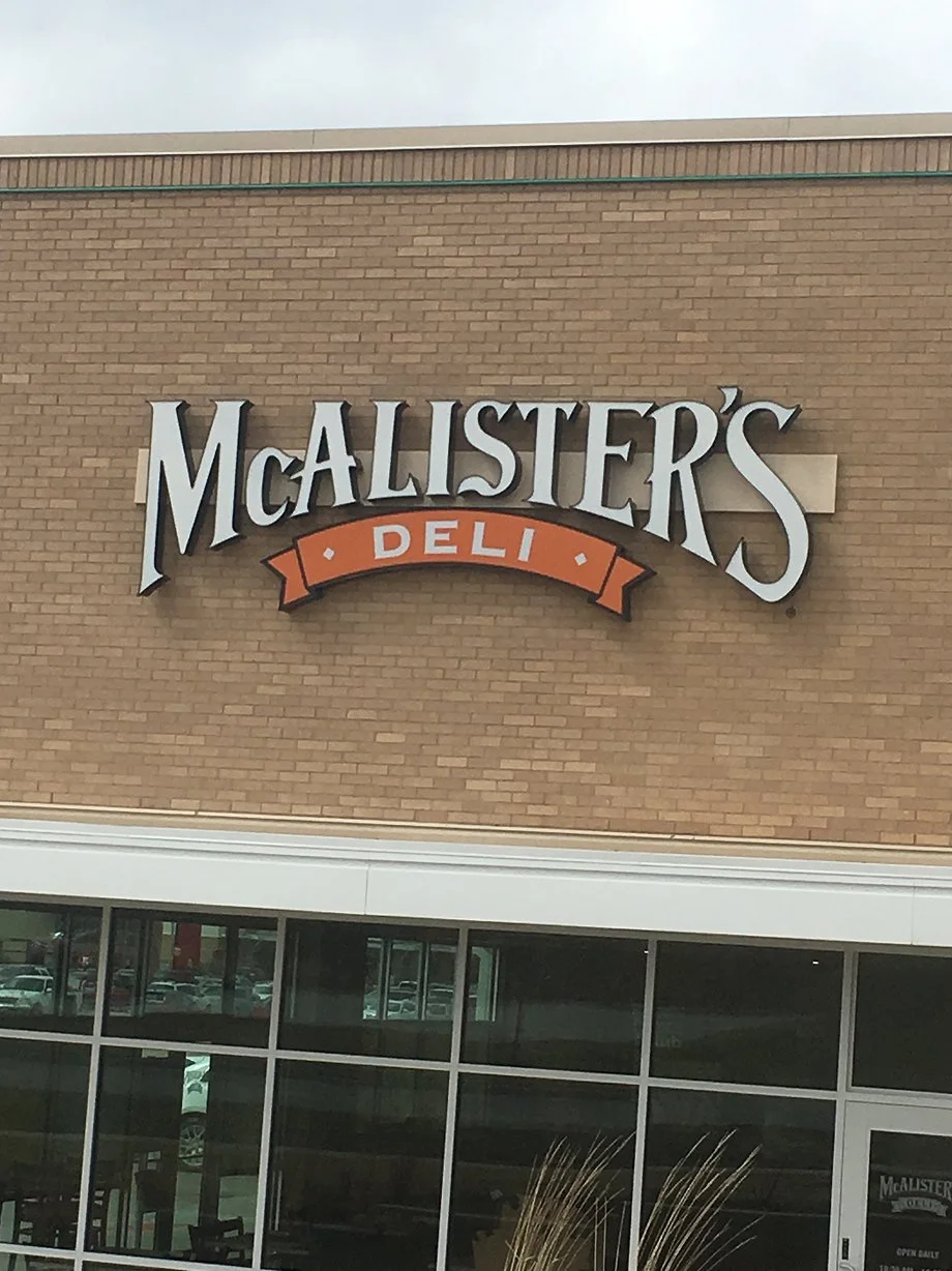 Exterior of McAllister's Deli restaurant with a brick wall and large sign displaying the restaurant's name and logo.