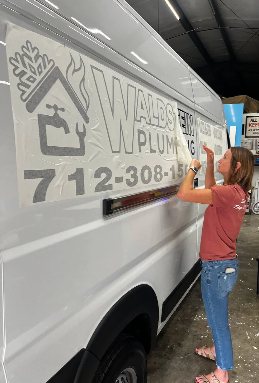 A woman is applying a vinyl decal to the side of a white service van with business information, including a phone number, for a plumbing company named WALS PLUMBING.