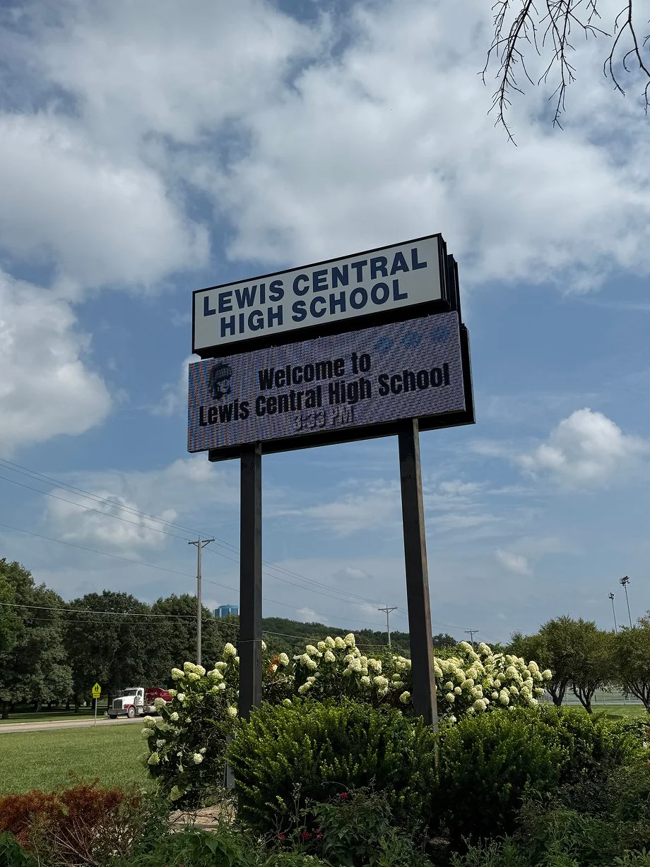 Sign for Lewis Central High School with a welcome message, surrounded by bushes and trees, under a partly cloudy sky.
