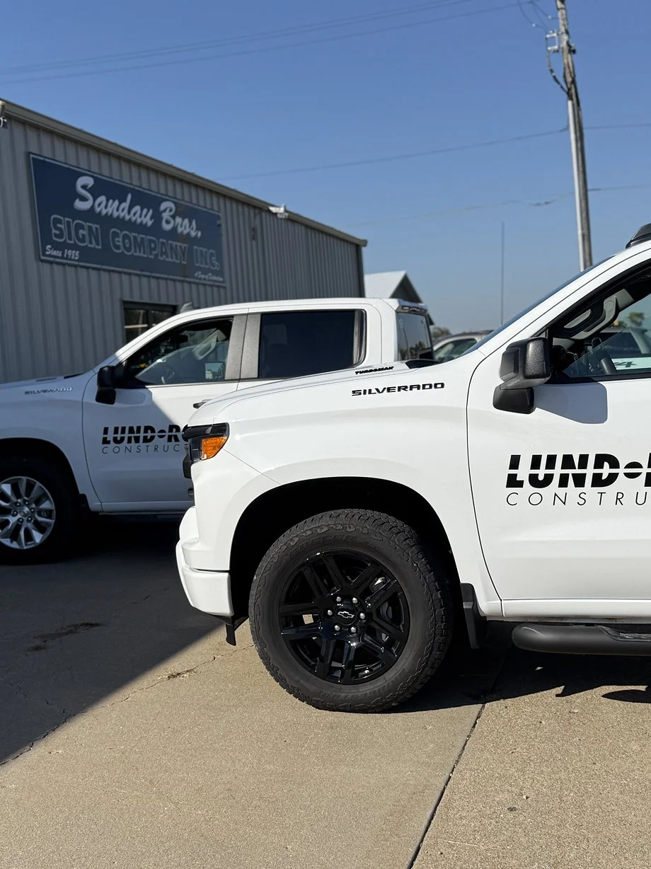 Two white pickup trucks with 'LUNDR' and 'CONSTRUCTION' logos parked in front of a building with a sign that reads 'Sandau Bros SIGN COMPANY INC.' under a blue sky.