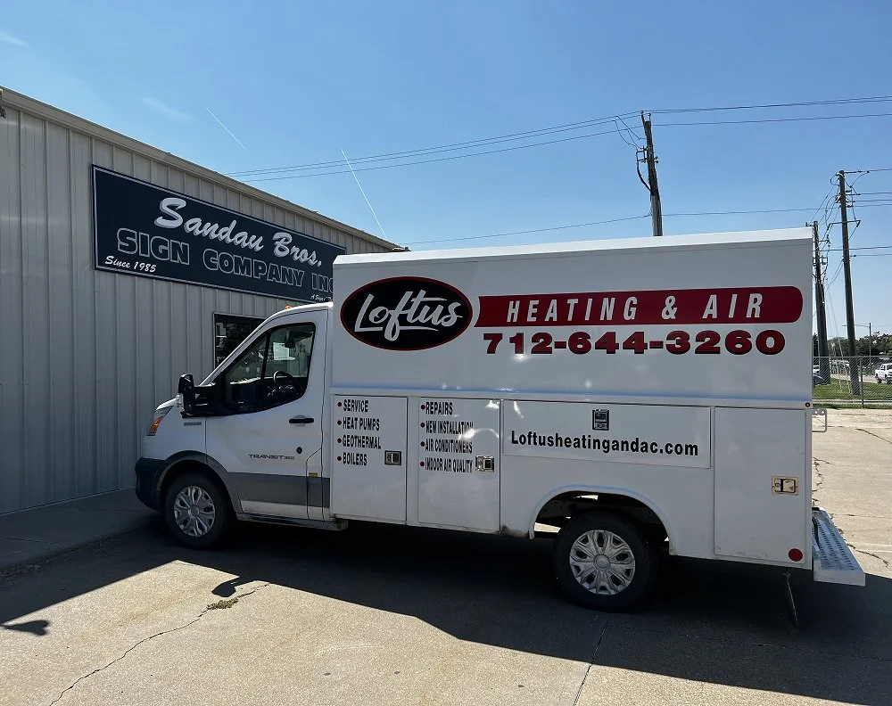 White service van for Loftus Heating & Air, parked outside a building with a sign that reads Sandau Bros Sign Company Inc.