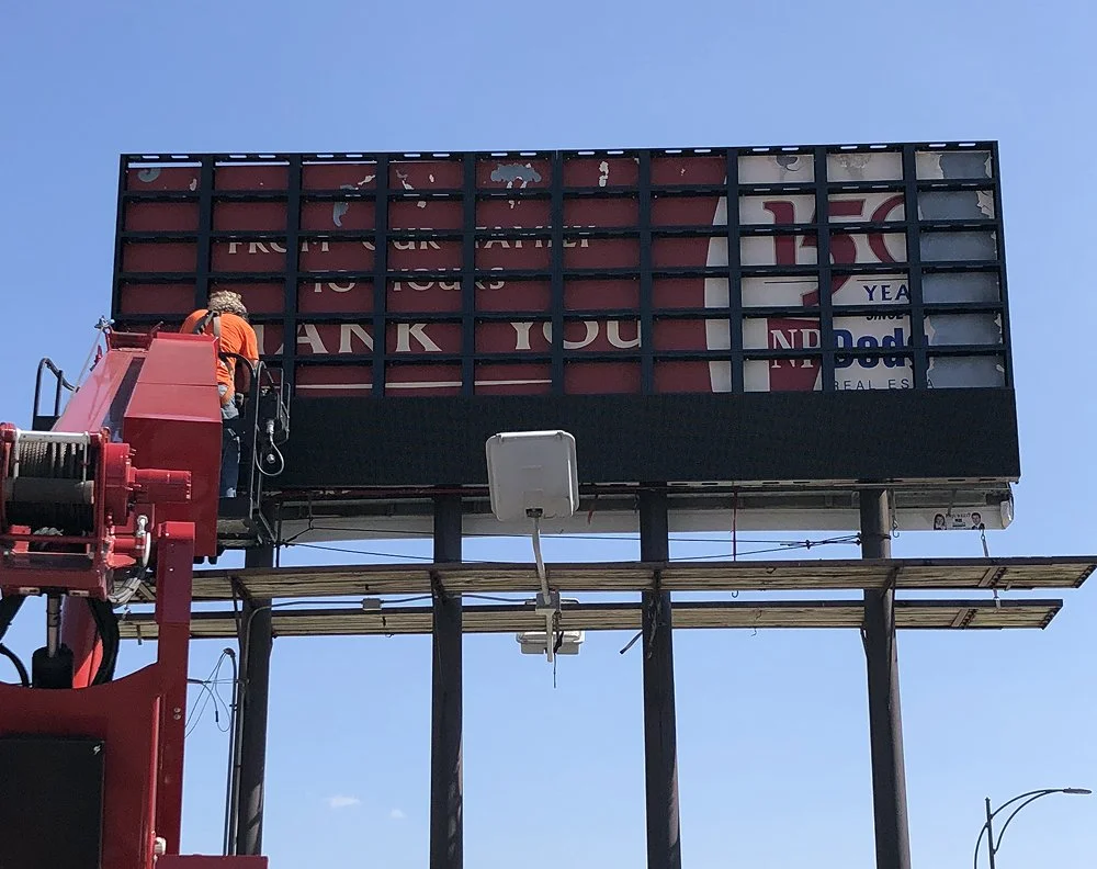 A worker in an orange shirt on a lift cleaning or repairing an outdoor signboard against a clear blue sky.