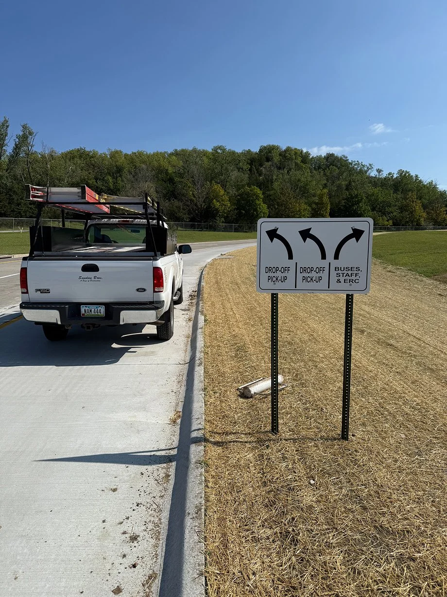 A white pickup truck parked on the side of a road next to a sign with directions for drop-off pickup and buses, staff, and ERC. The sign has three arrows pointing left, straight, and right. The area is grassy with trees on a hill in the background an