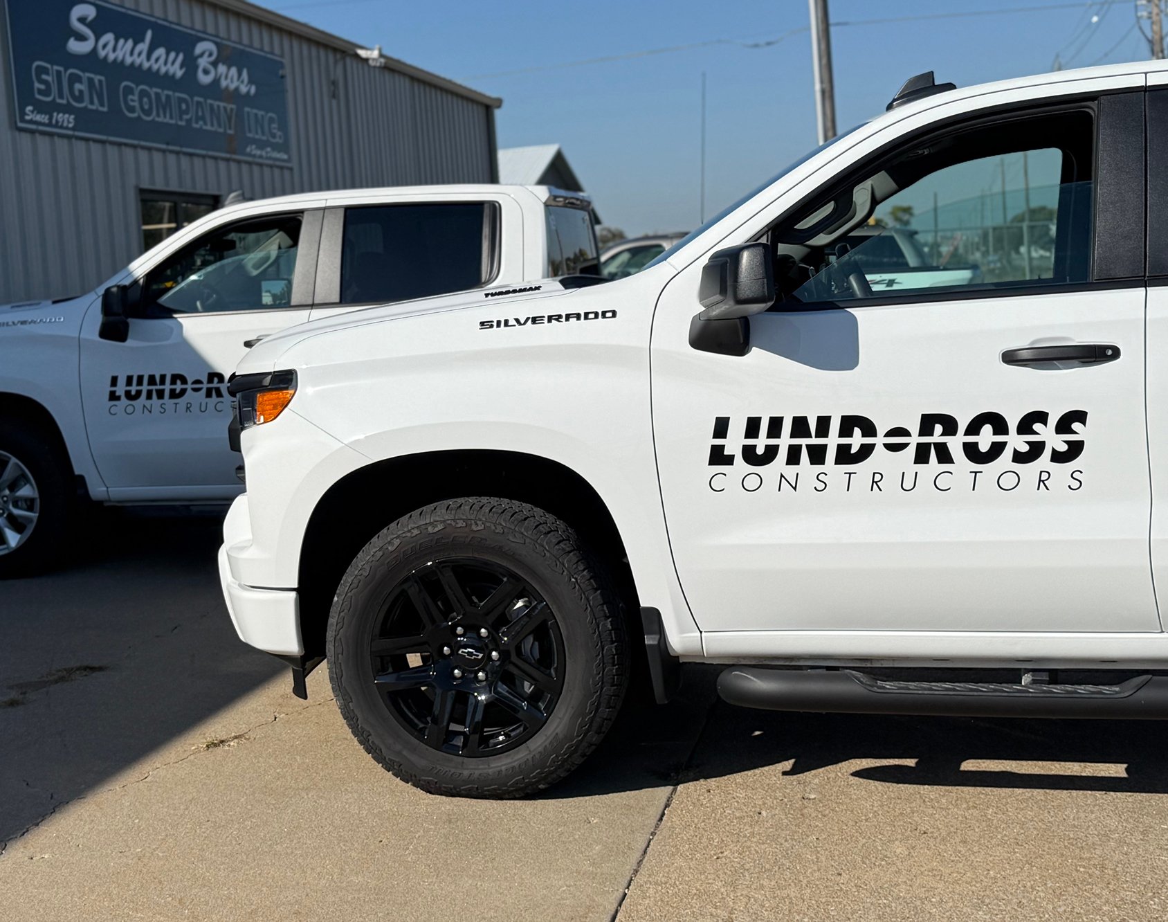 Two white pickup trucks with 'LUNDROSS CONSTRUCTORS' logos parked on a concrete lot, with one truck in the foreground and another slightly behind, and a sign for a sign company building in the background.