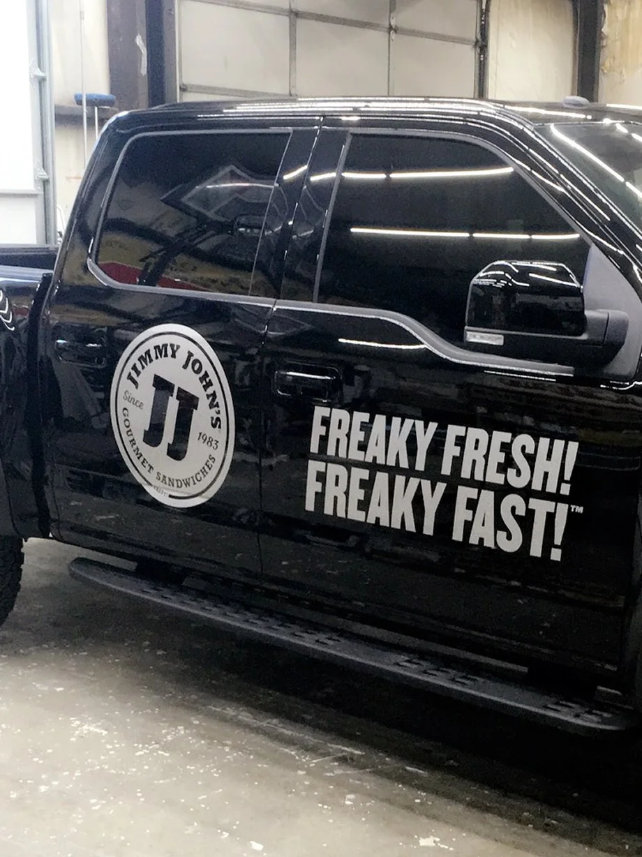 Black pickup truck with a Jimmy John's logo and slogan 'Freaky Fresh! Freaky Fast!' on the side, parked inside a garage.
