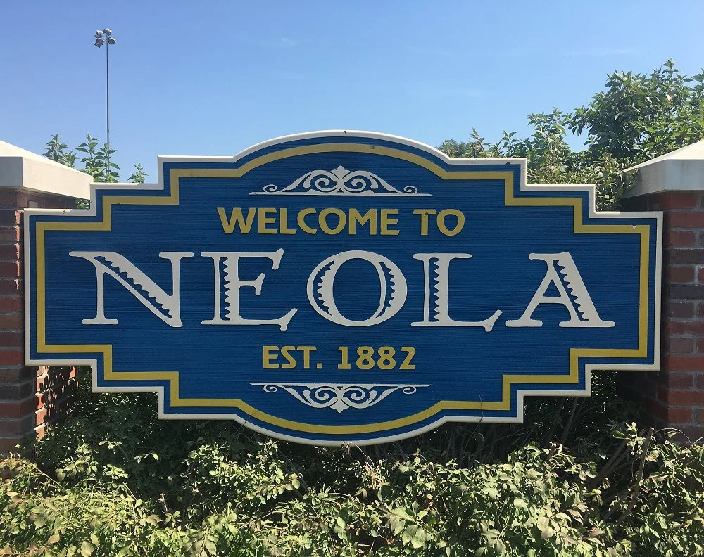 Welcome sign for Neola, established in 1882, with decorative elements and nearby greenery.