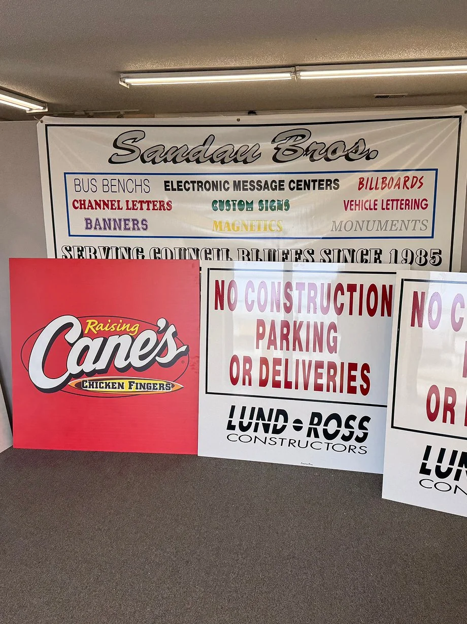 Signs in a business office for Sandau Bros and Lund-Ross Constructors, including one with rules about no construction parking or deliveries, and another promoting Raising Cane's Chicken Fingers.