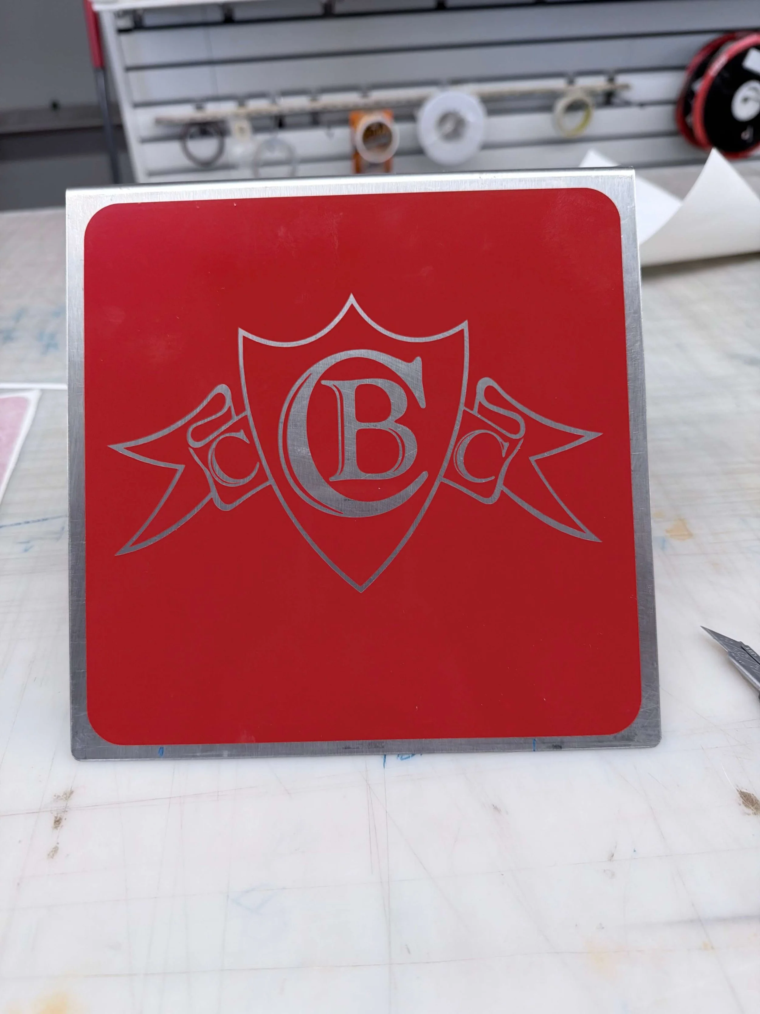 Red square sign with a silver crest featuring a large letter B in the center, flanked by the letters C and A on ribbons on either side.