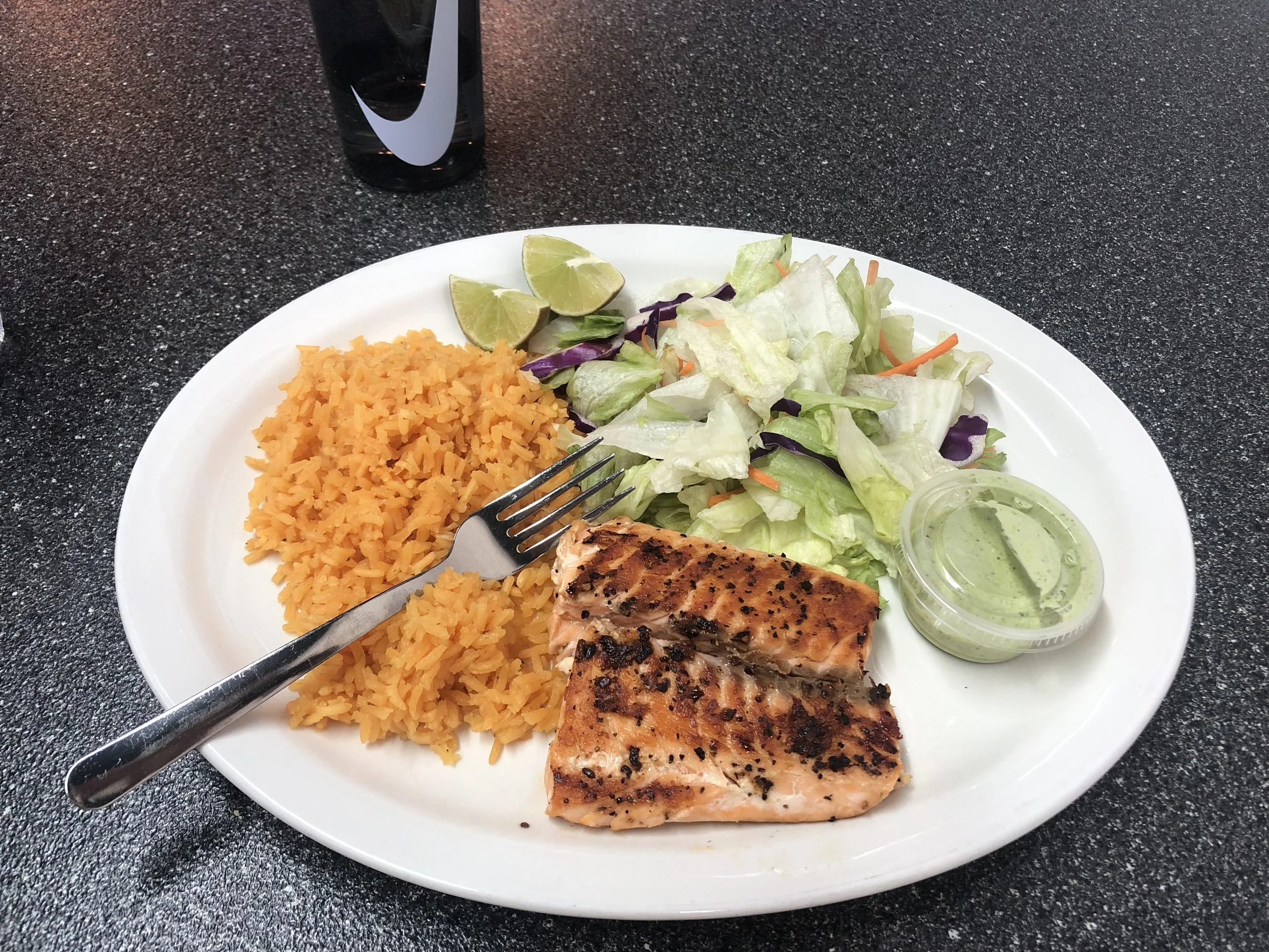 Grilled salmon fillet with rice, salad, lime wedges, and green dipping sauce on a white plate.
