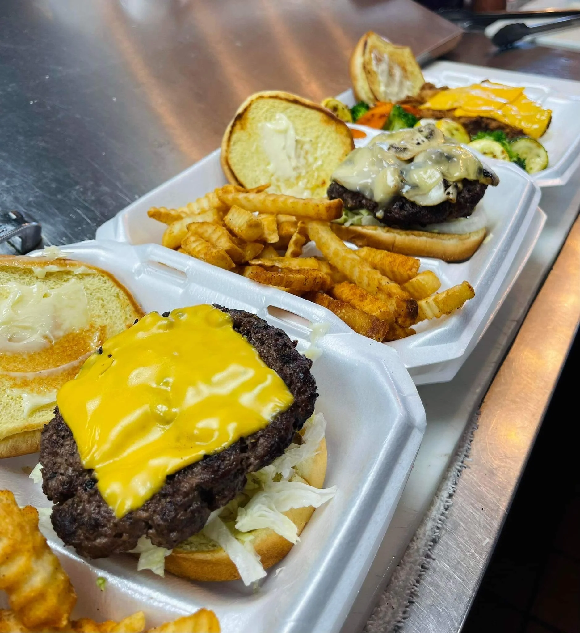 Two styrofoam trays with cheeseburgers, fries, and vegetables on a stainless steel counter.