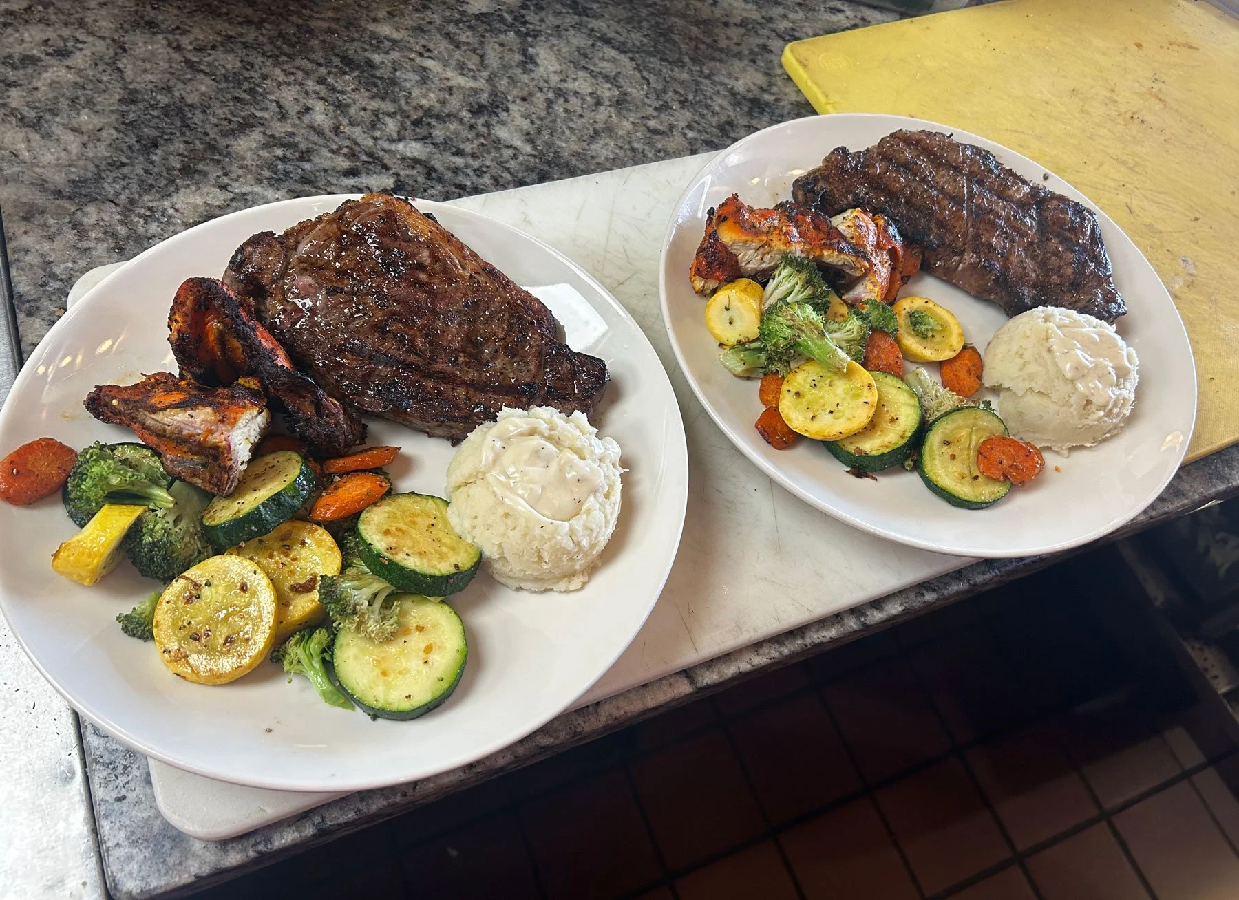 Two plates of grilled steak with roasted vegetables, mashed potatoes, and gravy, on a kitchen counter.